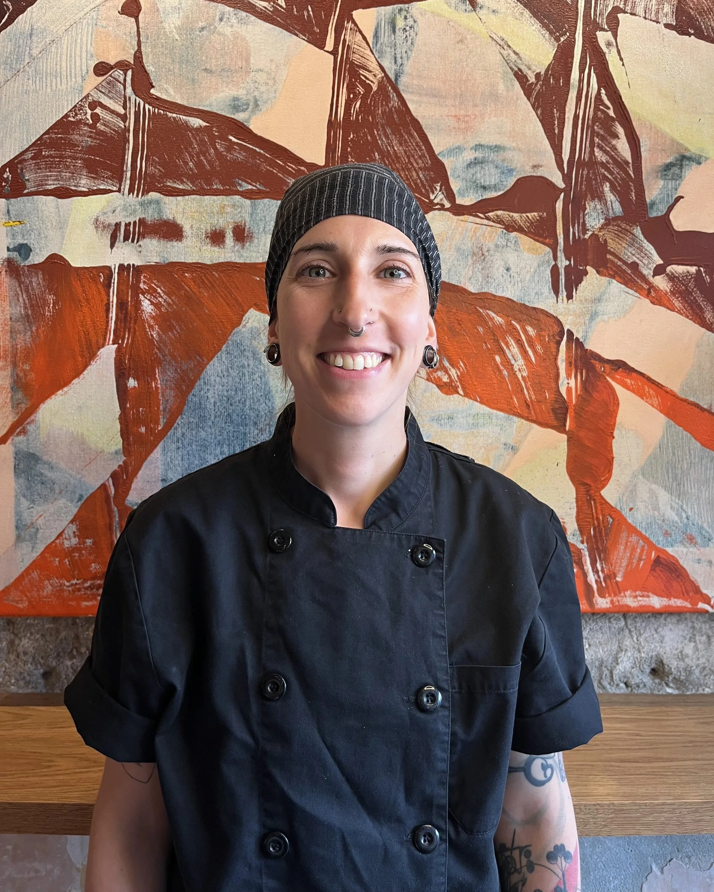 A woman with a black and gray striped head covering, smiling, wearing a black chef's coat with large buttons, standing in front of a colorful abstract painting with shades of orange, brown, and beige. The background includes a wooden surface and a textured wall.
