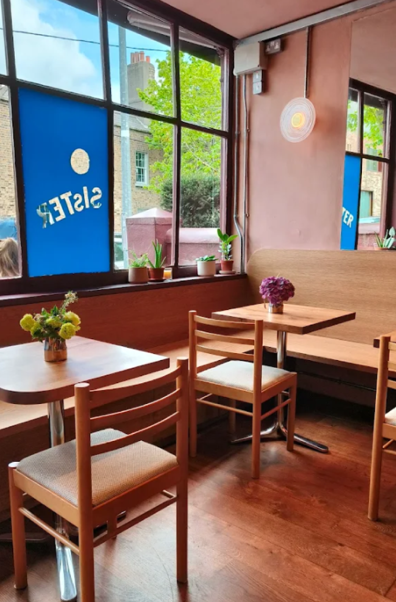 Cozy corner of a cafe with wooden tables and chairs, decorated with potted plants and flowers, large windows letting in natural light, and a sign on the window that reads 'SILSTEK'.