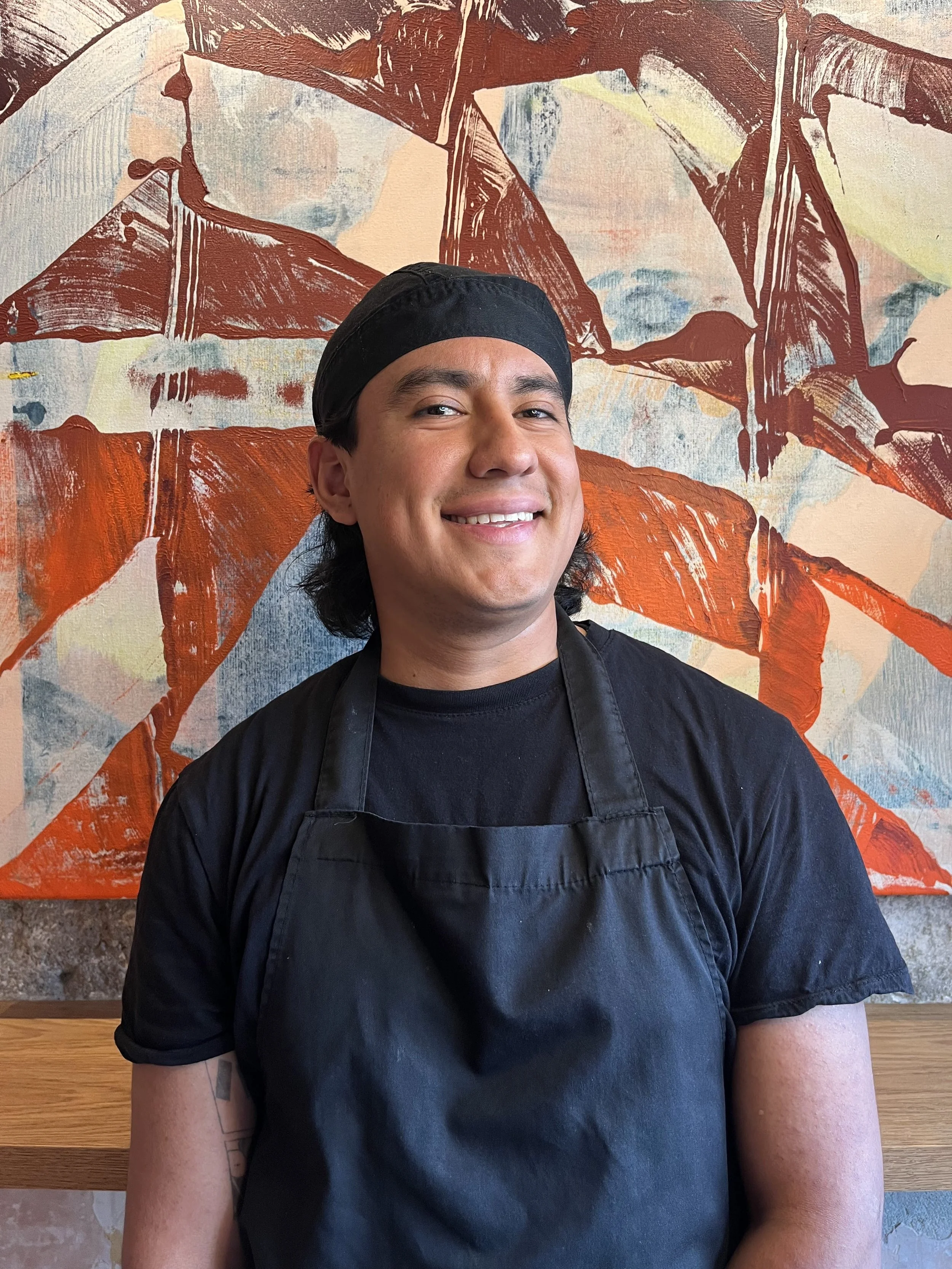 A smiling man wearing a black chef's hat and black apron, standing in front of a colorful abstract mural with red, beige, blue, and yellow hues.