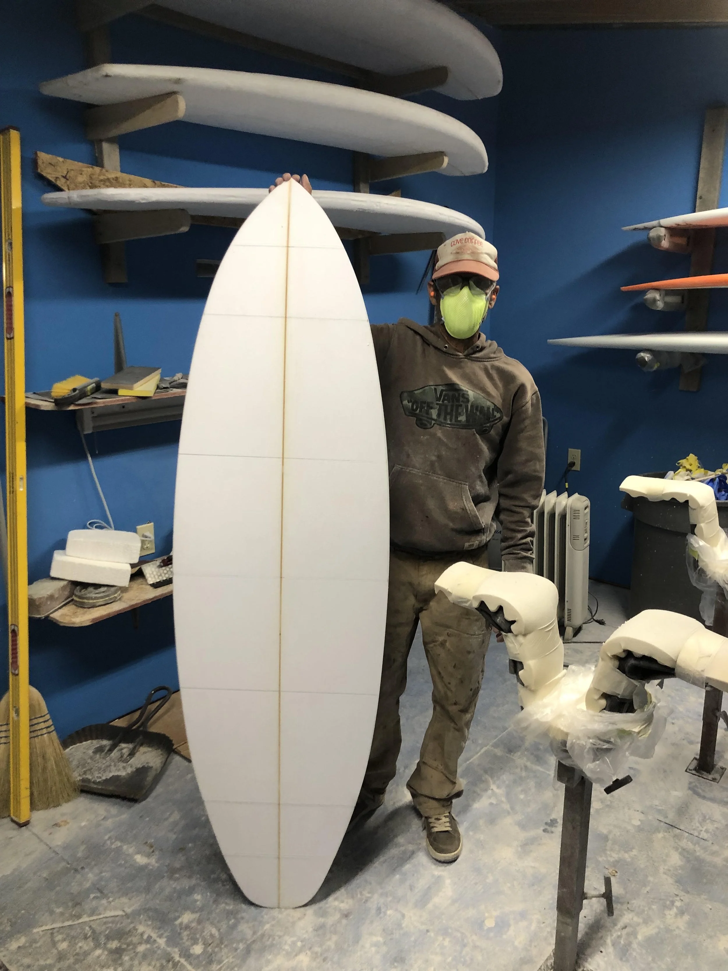 Person wearing a mask and cap standing next to a surfboard in a workshop, surrounded by surfboard blanks, tools, and equipment.