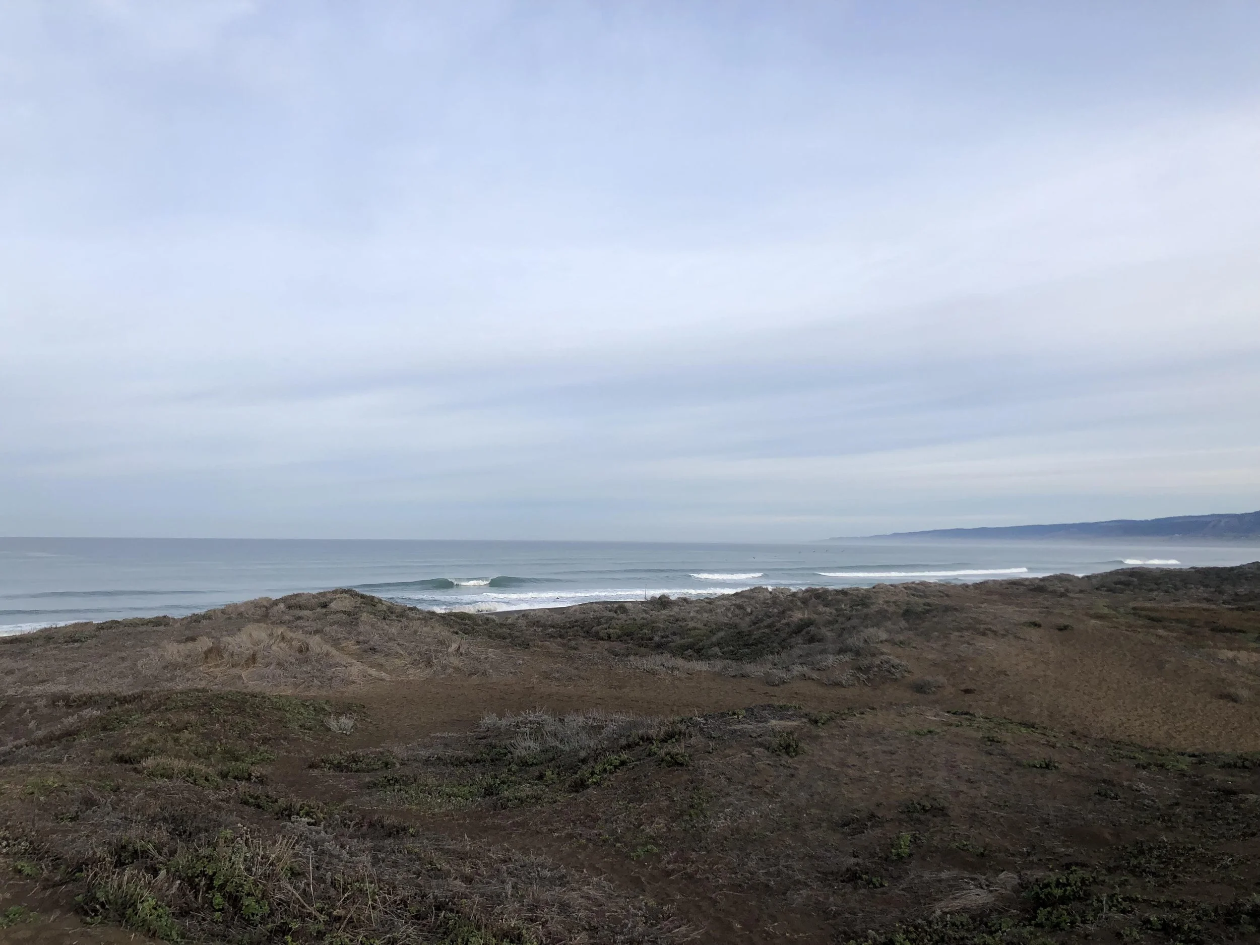 Photo of a coastal landscape showing the ocean, rolling waves, a sandy beach, and cloudy sky.