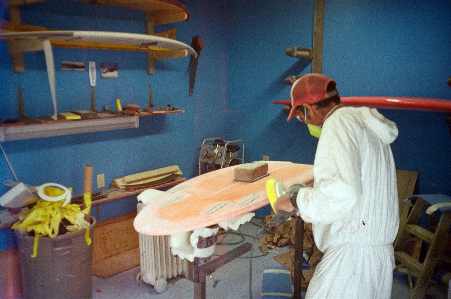 A person wearing protective gear, including gloves, mask, and cap, shapes a surfboard in a workshop with blue walls and various tools on the shelves.