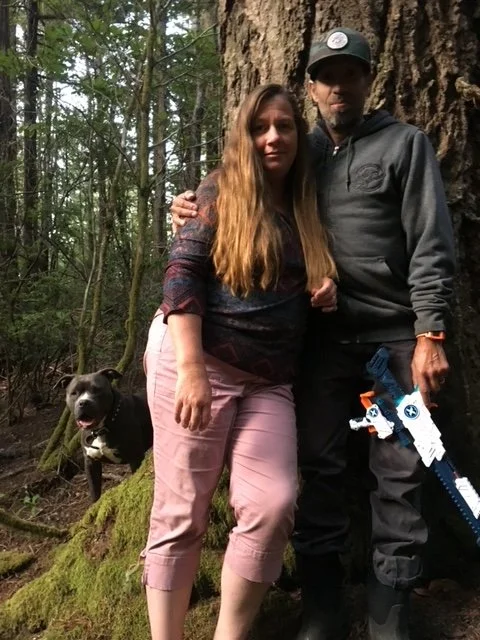 A woman and a man standing close together in a forest, with a black dog sitting nearby on a moss-covered log.