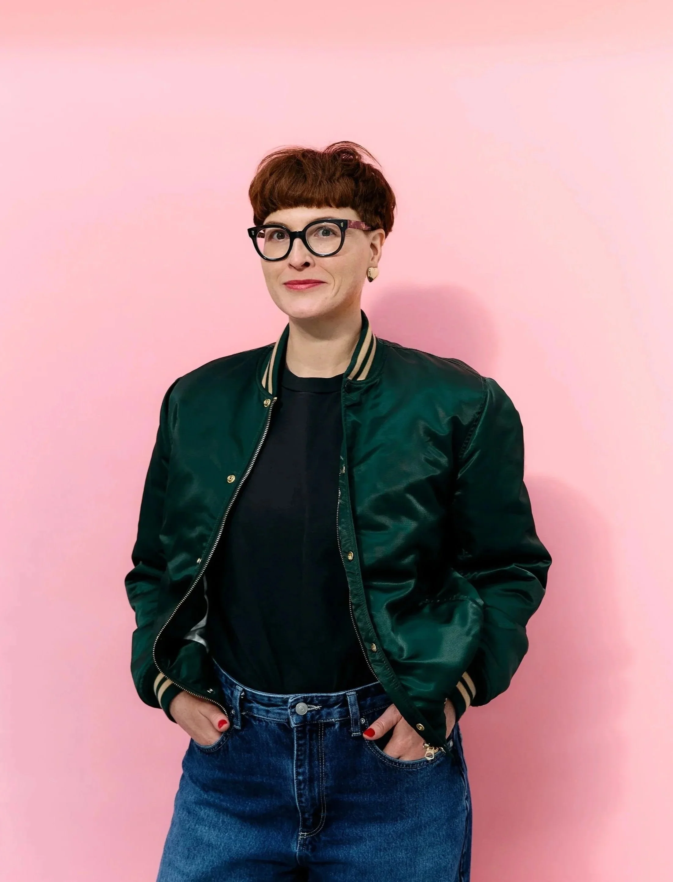 Frau mit rotem Bob, schwarzen Brillen, grüner Bomberjacke und Jeans vor rosa Hintergrund.