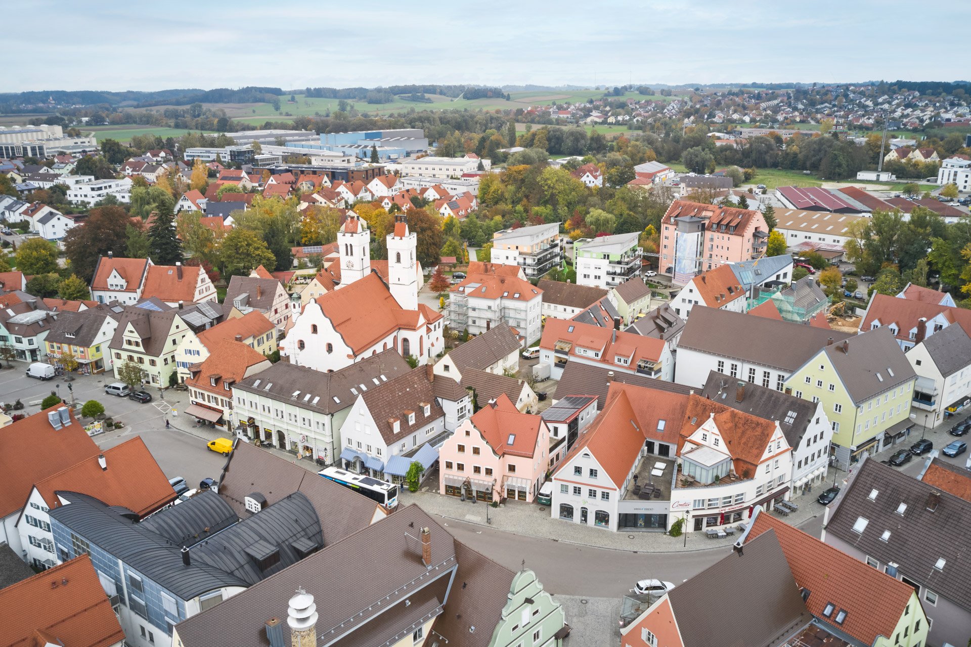 Luftaufnahme einer kleinen Stadt mit roten und schwarzen Ziegeldächern, Kirchen mit weißen Türmen und bunten Gebäuden im Vordergrund, umgeben von grünen Bäumen und offenen Feldern im Hintergrund.