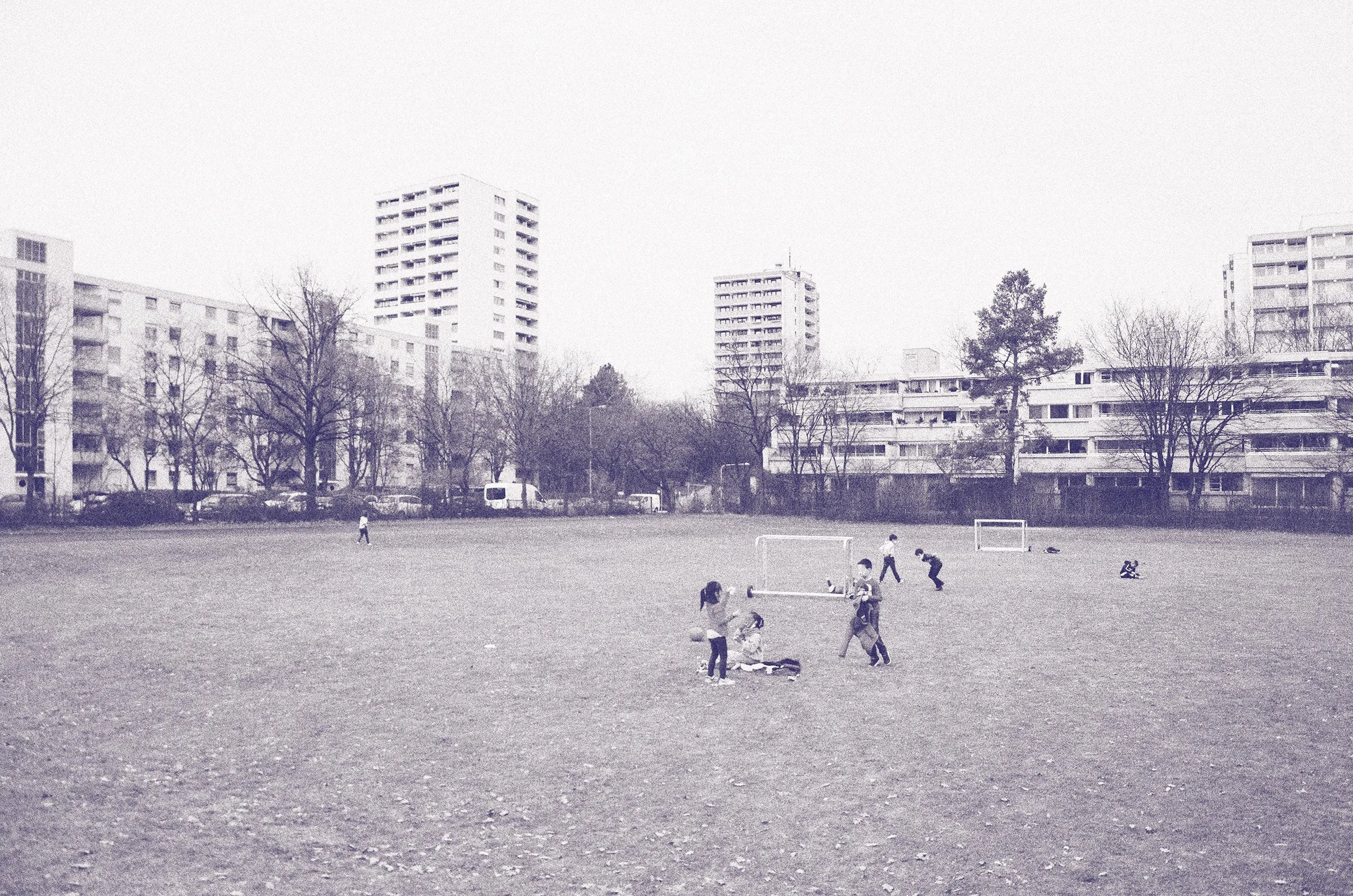 Kinder spielen Fußball auf einem Sportplatz, im Hintergrund moderne Hochhäuser und Bäume, bei bewölktem Himmel.