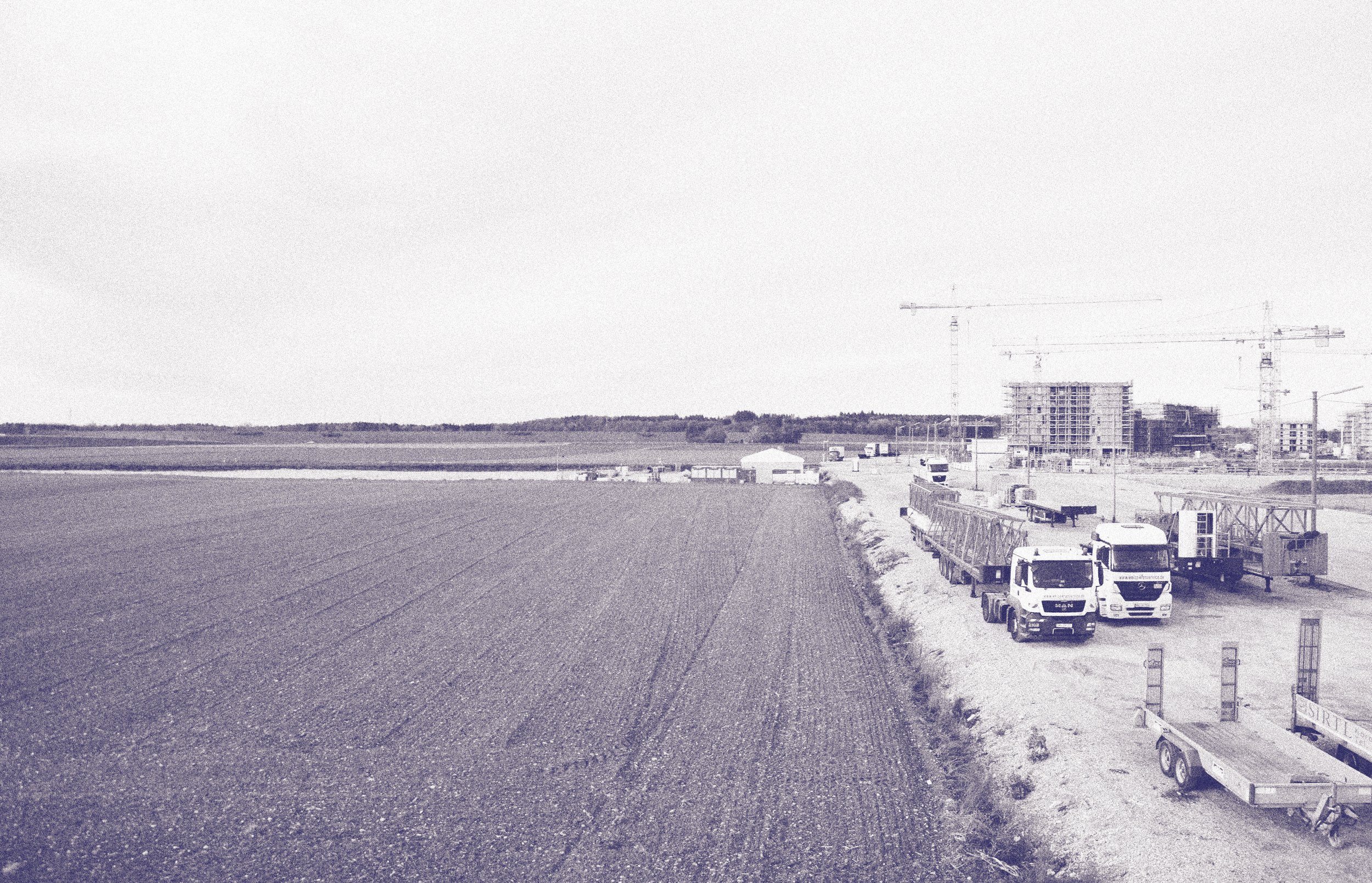 Bauernhof mit landwirtschaftlichen Feldern im Vordergrund und Baustelle mit Gebäudekonstruktionen und Kränen im Hintergrund in einer ländlichen Umgebung.