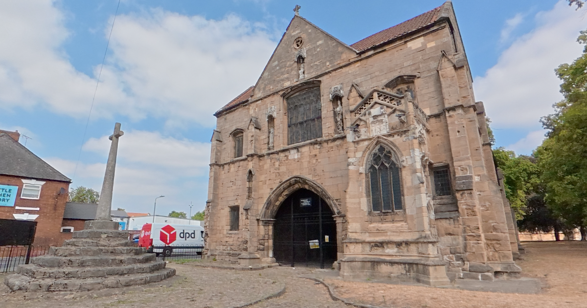 An old stone church with a pointed roof, gothic windows, and an arched entrance, situated in an outdoor area with trees and a cross monument nearby.
