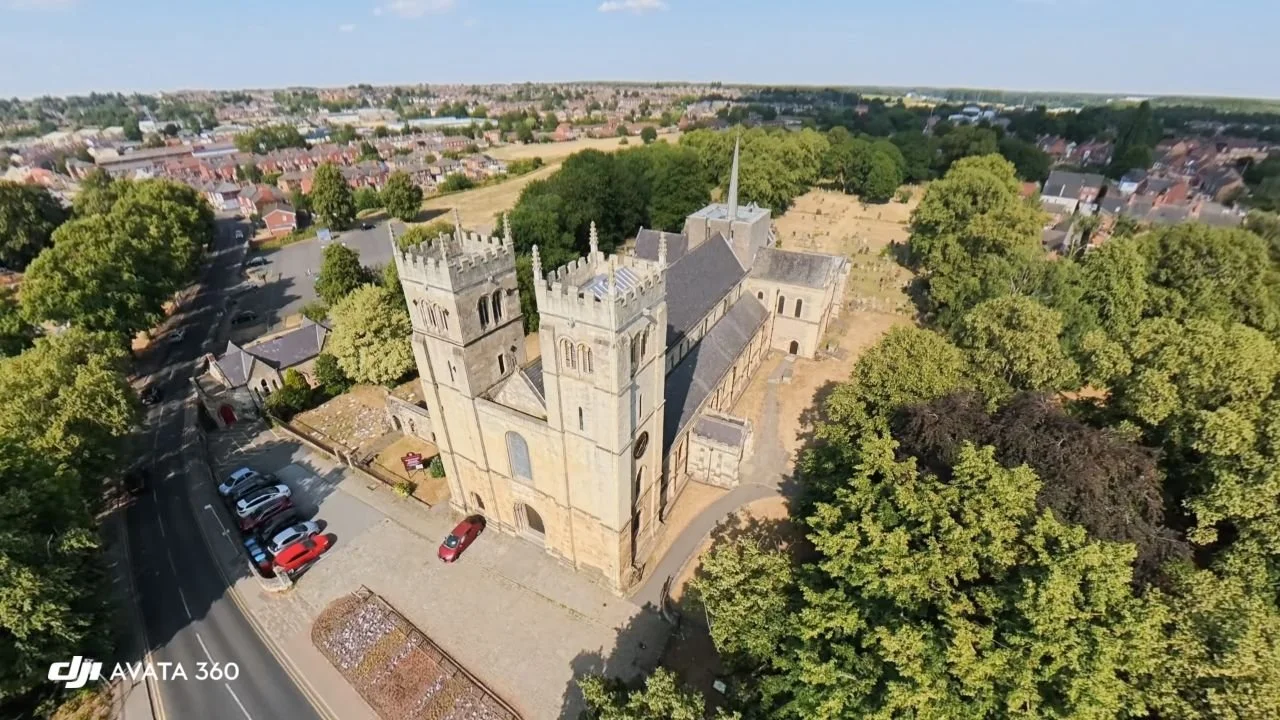 Aerial view of a historic stone church with gothic architecture, surrounded by trees and a small parking lot, in a suburban area.