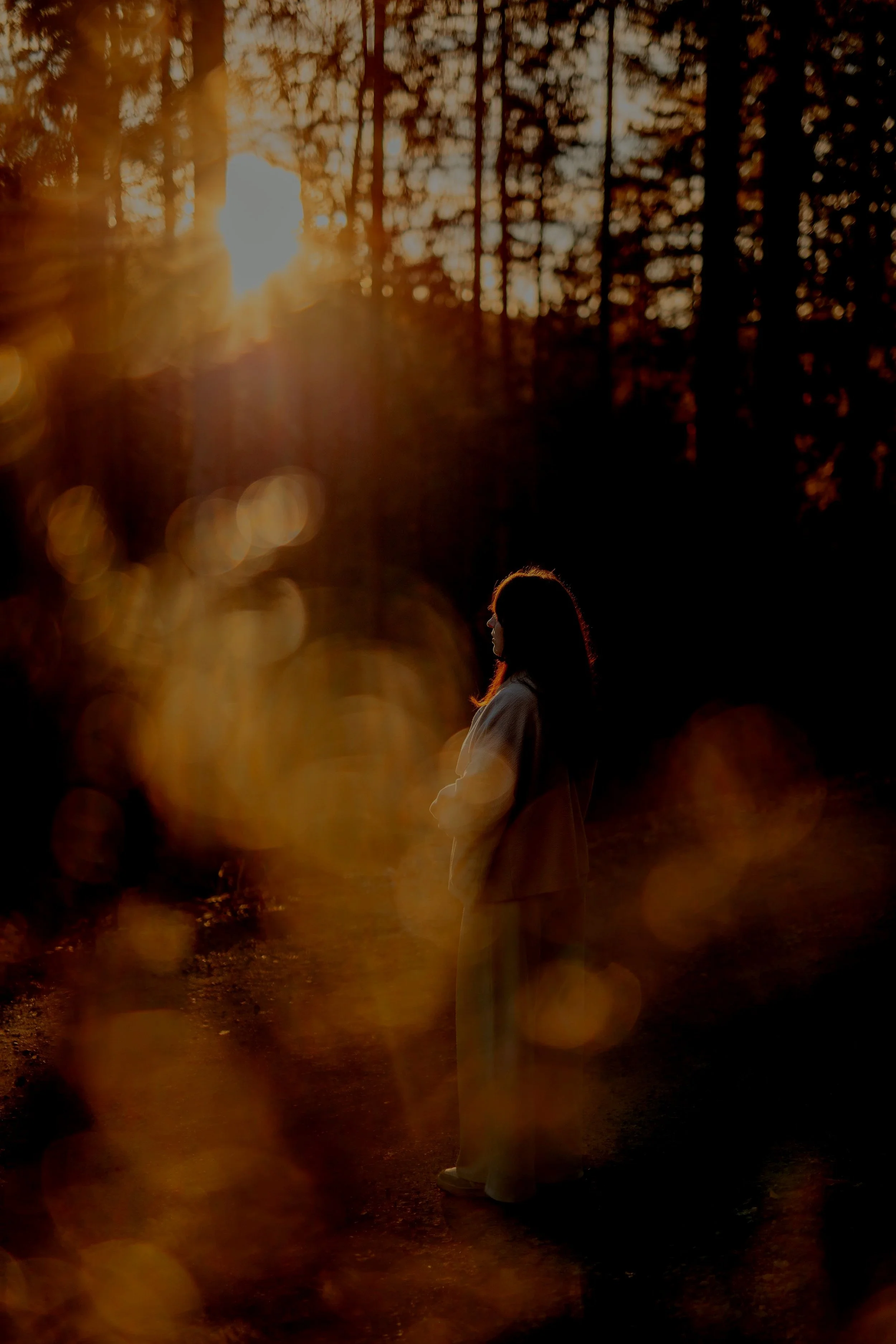 Eine Frau steht in einem dunklen Wald bei Sonnenuntergang, ihr Blick ist zur Seite gerichtet, im Hintergrund sind Bäume und das Sonnenlicht, das durch die Baumwipfel scheint.