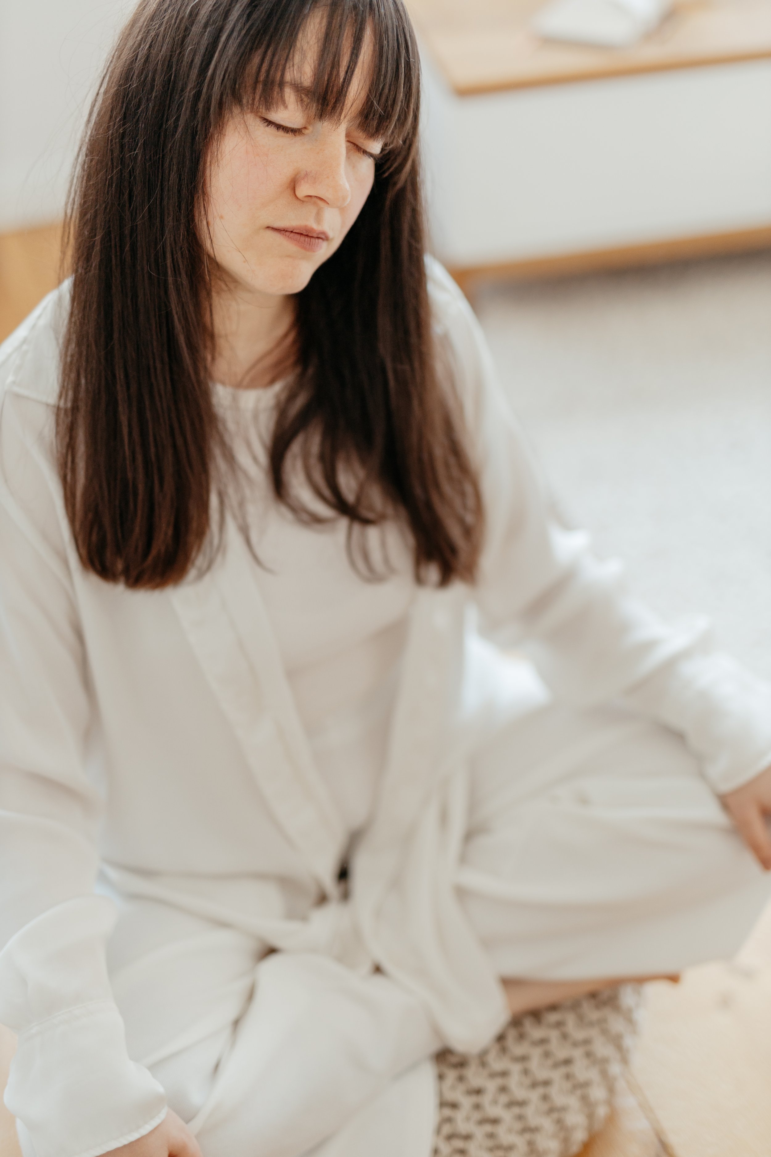 Eine Frau mit braunen, glatten Haaren, die ihre Augen geschlossen hat, sitzt in einem hellen Raum, trägt ein weißes Hemd und weiße Hosen, mit einem Teppich im Hintergrund.