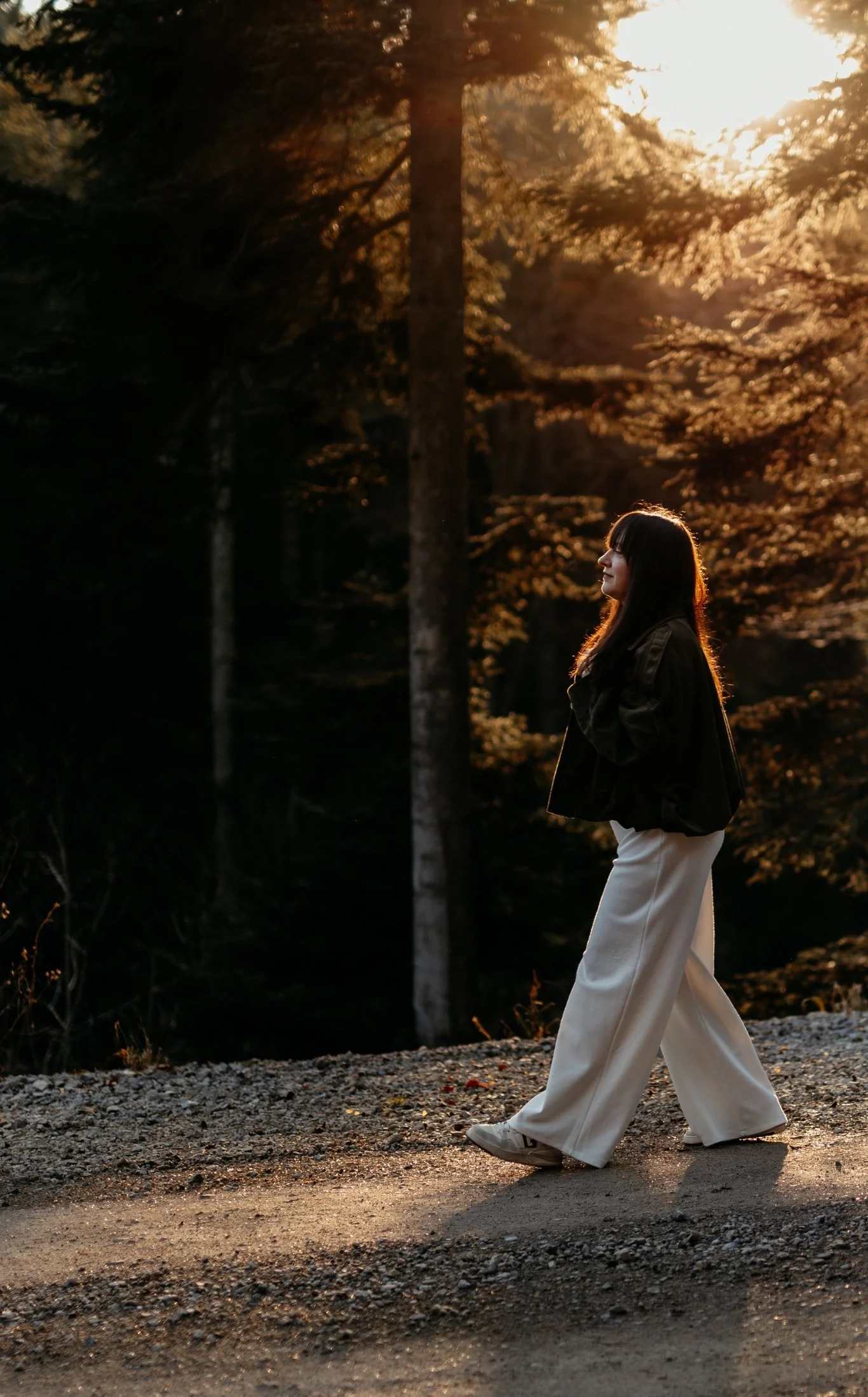 Junge Frau läuft in einem dunklen Wald bei Sonnenuntergang, trägt weiße Hosen und eine schwarze Jacke.