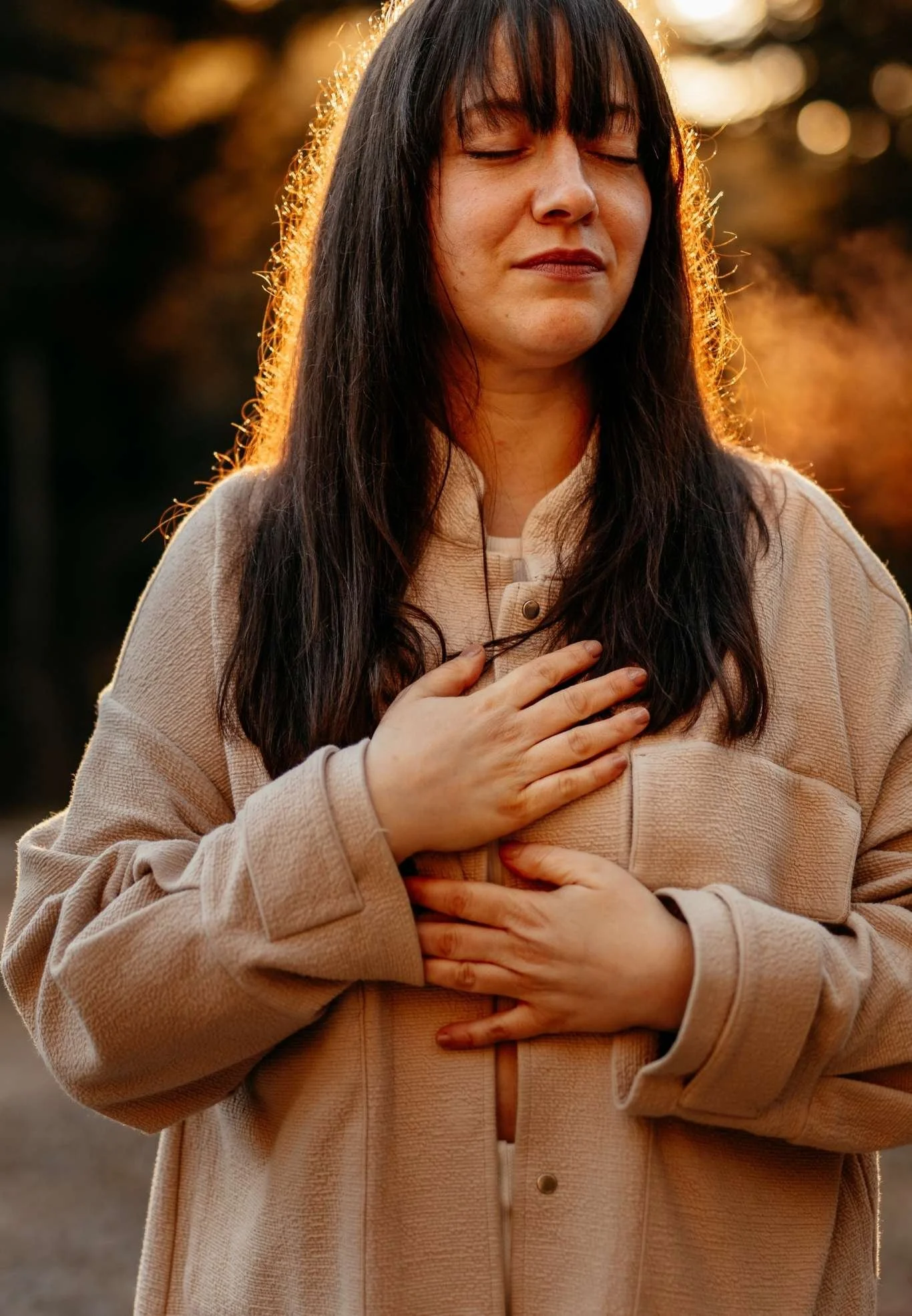 Eine Frau mit langem dunklem Haar, die die Augen geschlossen hat und die Hand auf die Brust legt, während sie draußen bei Sonnenuntergang oder Sonnenaufgang steht. Sie trägt einen beige Mantel.