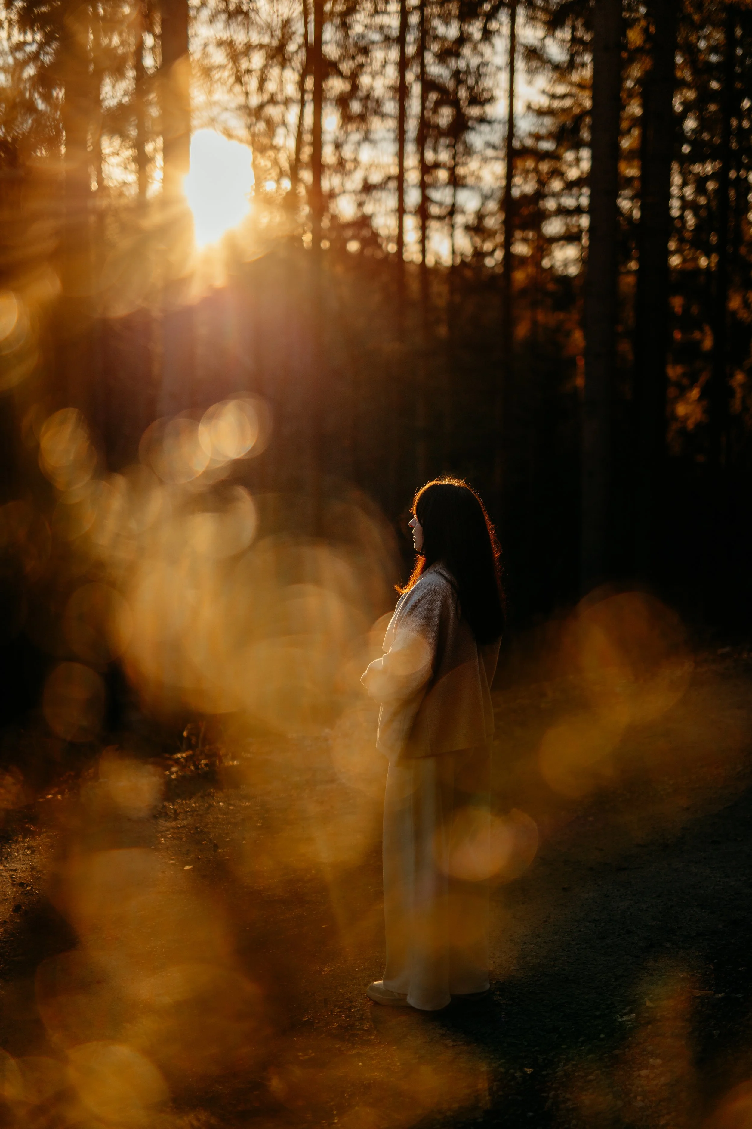 Eine Frau steht in einem sonnendurchfluteten Wald im Sonnenuntergang, sichtbar nur von der Seite mit langen dunklen Haaren und weißer Kleidung.
