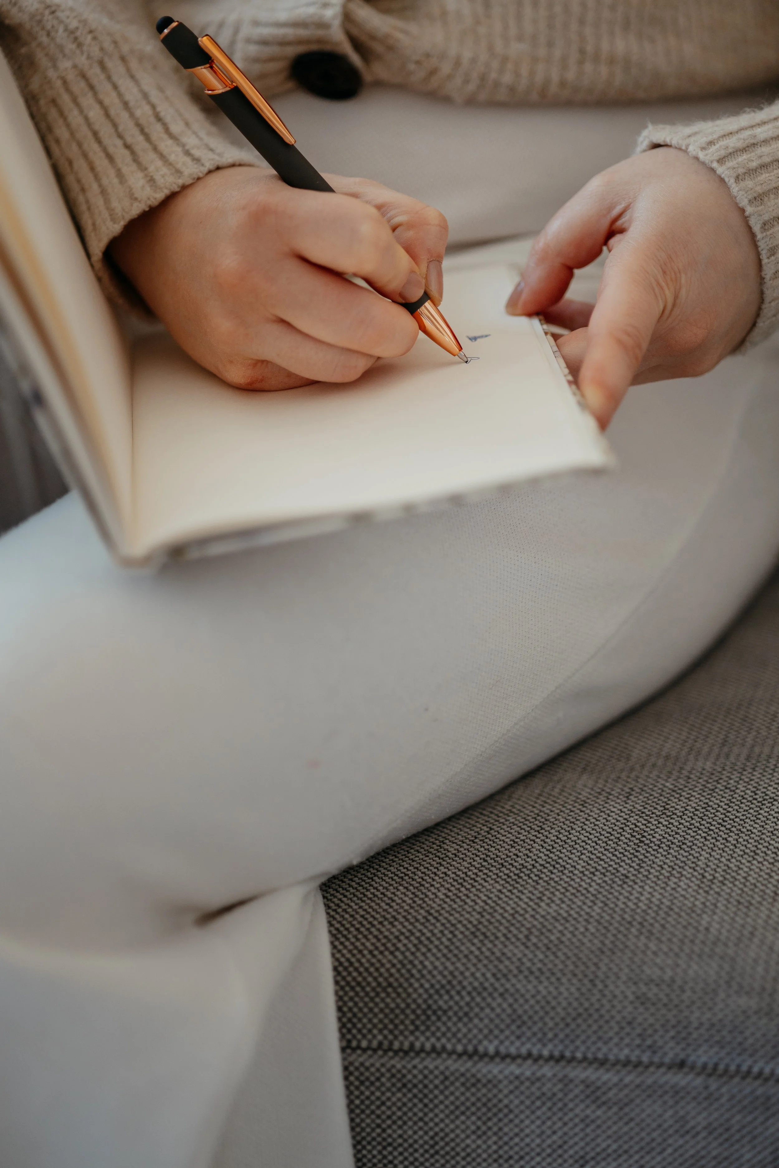 Person schreibt in ein Notizbuch mit einem schwarzen und kupferfarbenen Kugelschreiber, trägt einen beige Pullover, sitzend auf einem grauen Stoffsessel.
