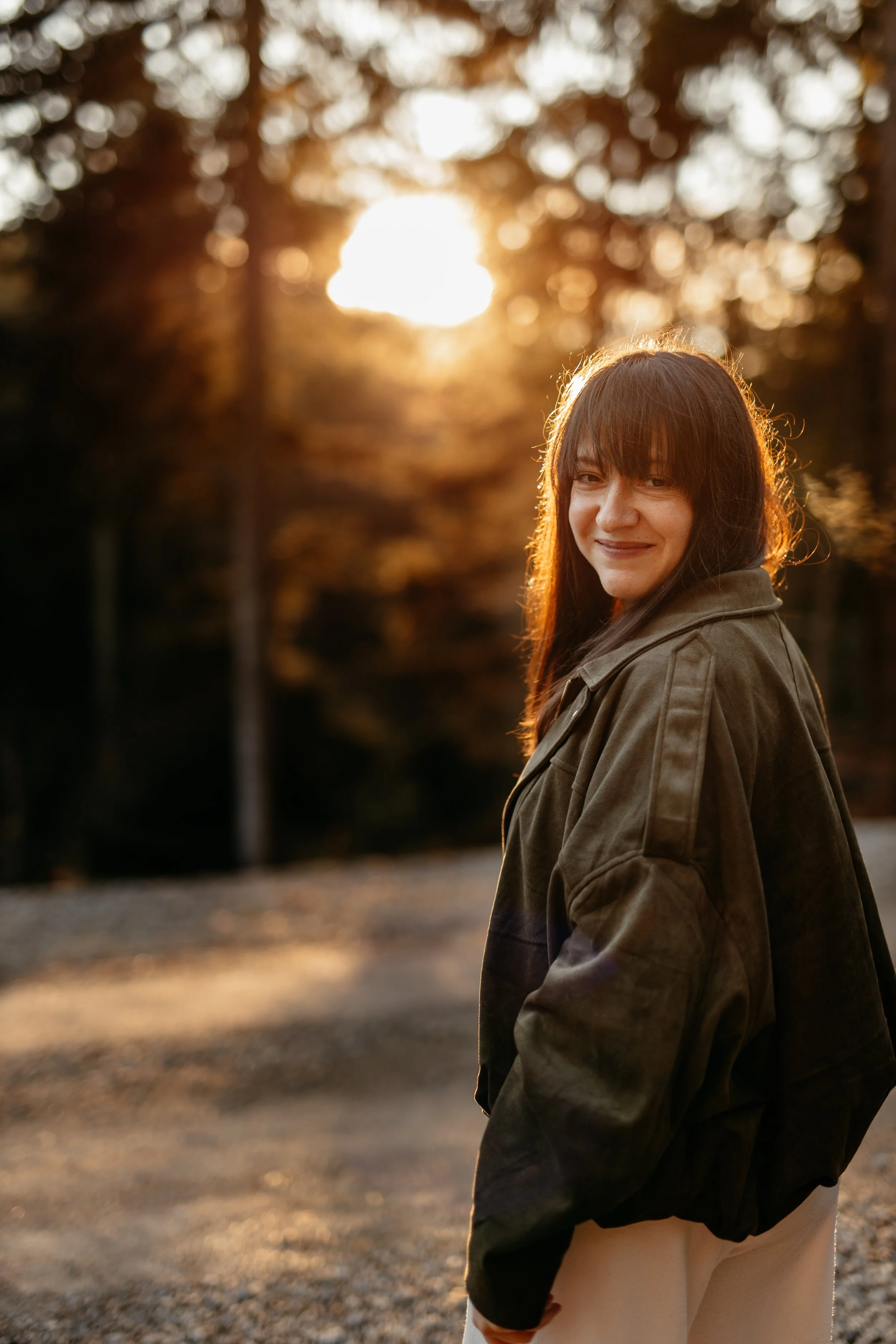 Ein lächelnder junger Mensch mit braunen Haaren und einem Jacke steht in einem Park bei Sonnenuntergang im Herbst, im Hintergrund sind Bäume mit goldenen Blättern.