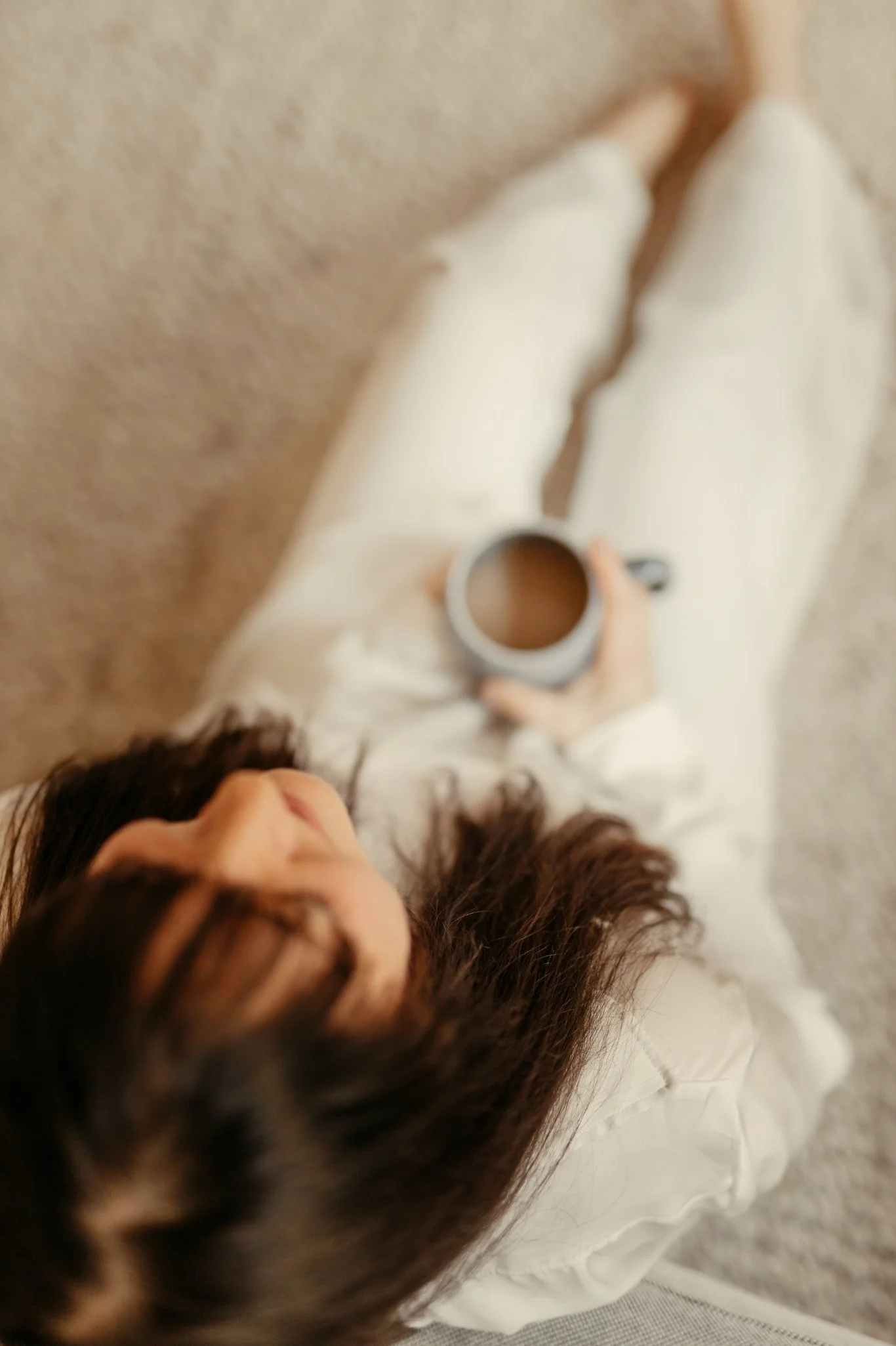 Eine Person liegt auf einer weißen Couch, hält eine Tasse Kaffee und trägt weiße Kleidung.