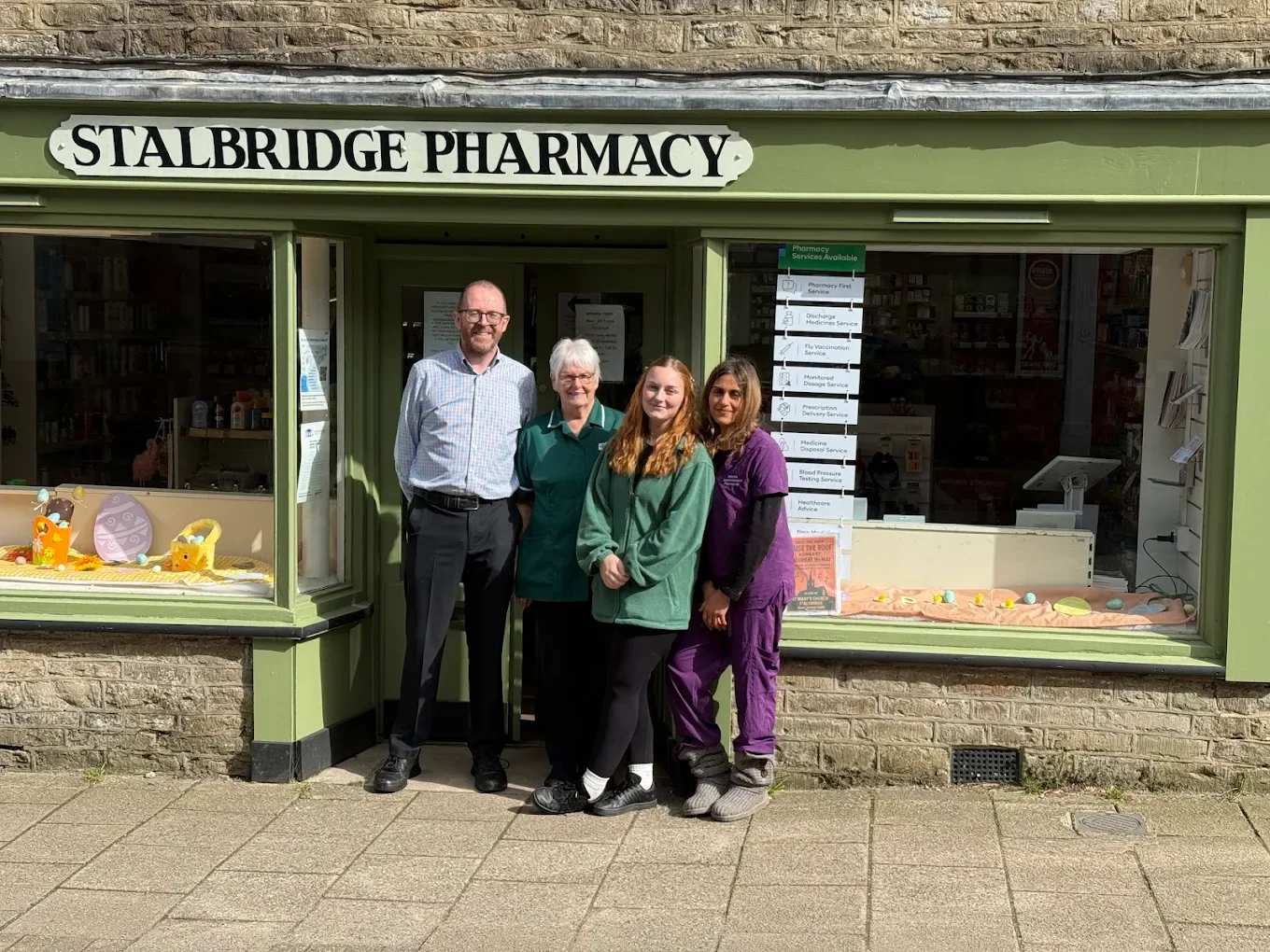 Four people standing outside the storefront of Stalbridge Pharmacy on a sunny day. The storefront has a green exterior with a large window display and a sign above that reads "Stalbridge Pharmacy." Inside the window are various products, and a sign with available pharmacy services is posted on the right side. The group includes two women in crew uniforms, one man in business casual, and one woman in purple scrubs. They are smiling and standing on a sidewalk.