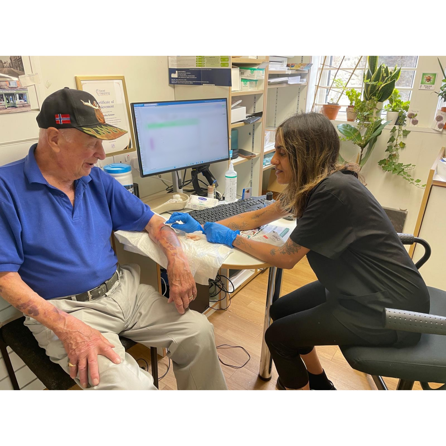 A woman in black scrubs providing a vaccination to an elderly man with a bandage on his arm, seated in a medical office with plants and medical supplies.