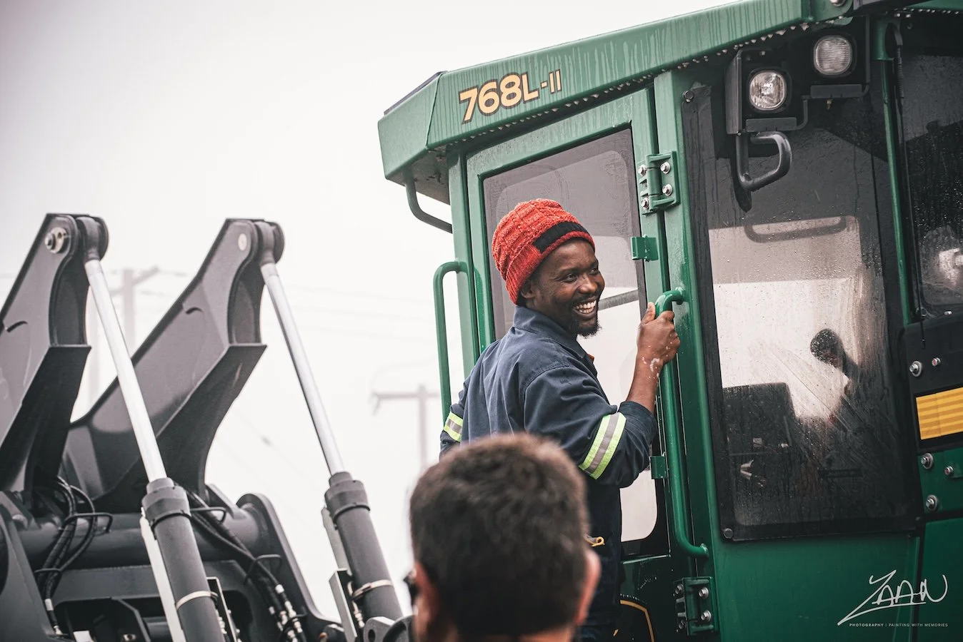 A smiling man with a red knit hat and work uniform standing inside a green construction vehicle, holding onto a door handle, with another person visible in the foreground.
