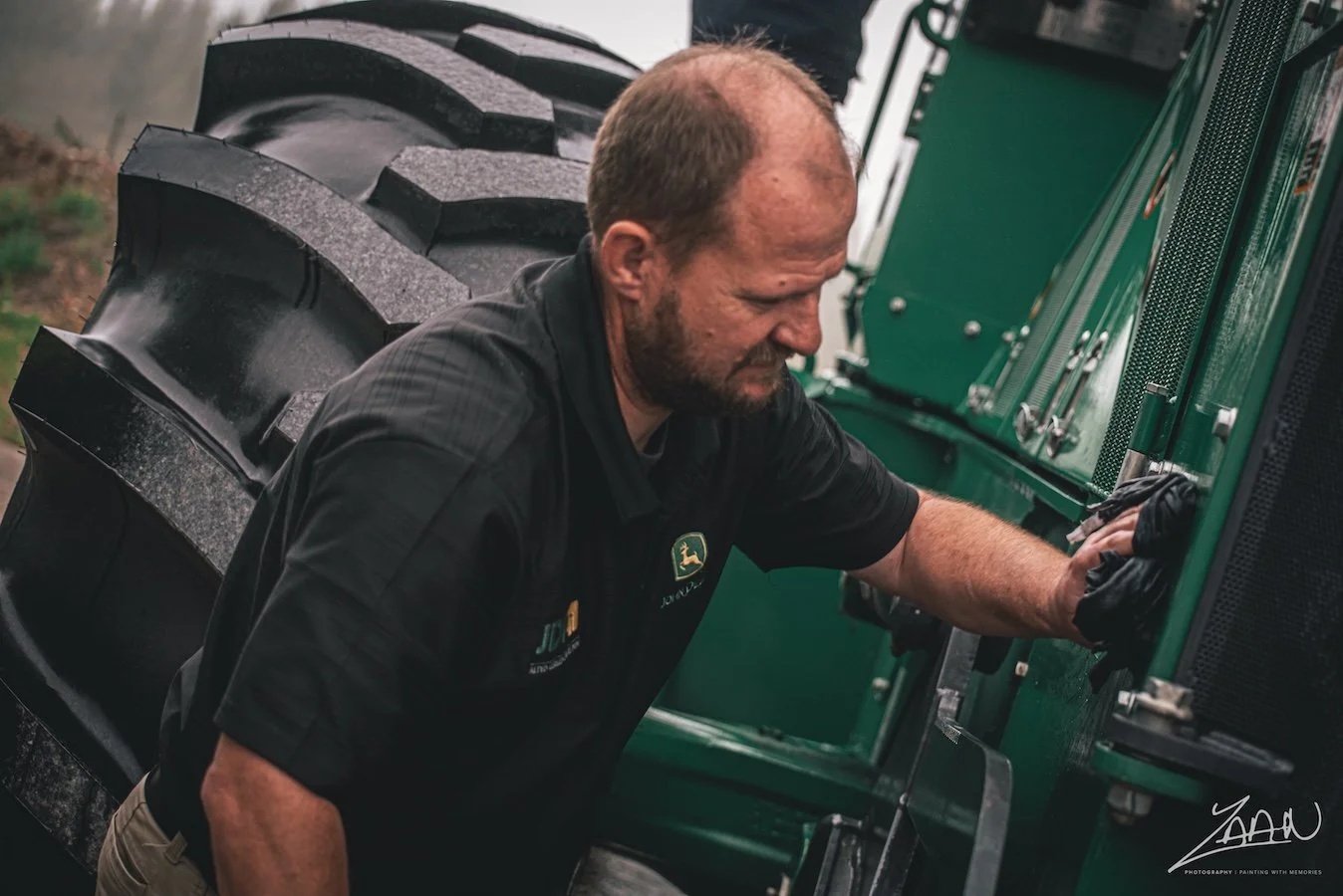 A man with a beard and short hair working on a green tractor. He is wearing a black polo shirt with a John Deere logo, black gloves, and is leaning over the side of the tractor, adjusting or inspecting something.