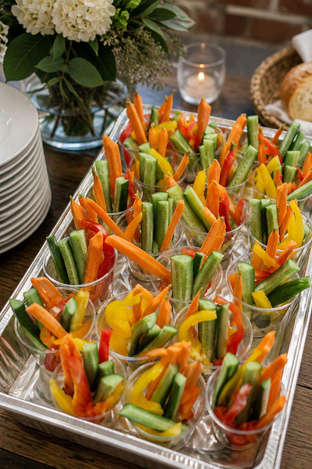 Tray of clear cups filled with cut vegetables, including cucumbers, carrots, and bell peppers, at a catered event or party.
