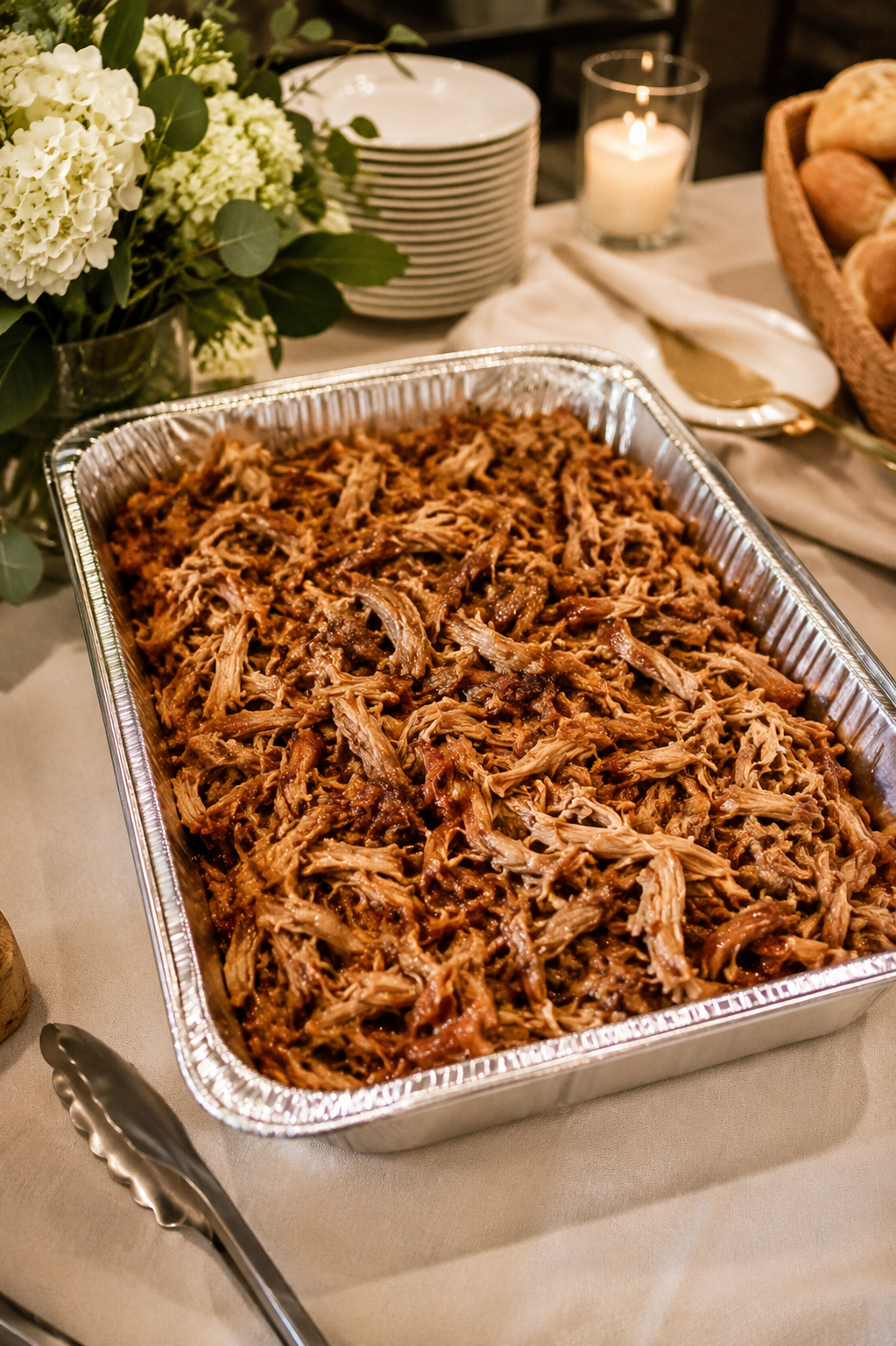 A tray of shredded barbecue pork served at a buffet table, with plates, a candle, flowers, and bread rolls in the background.