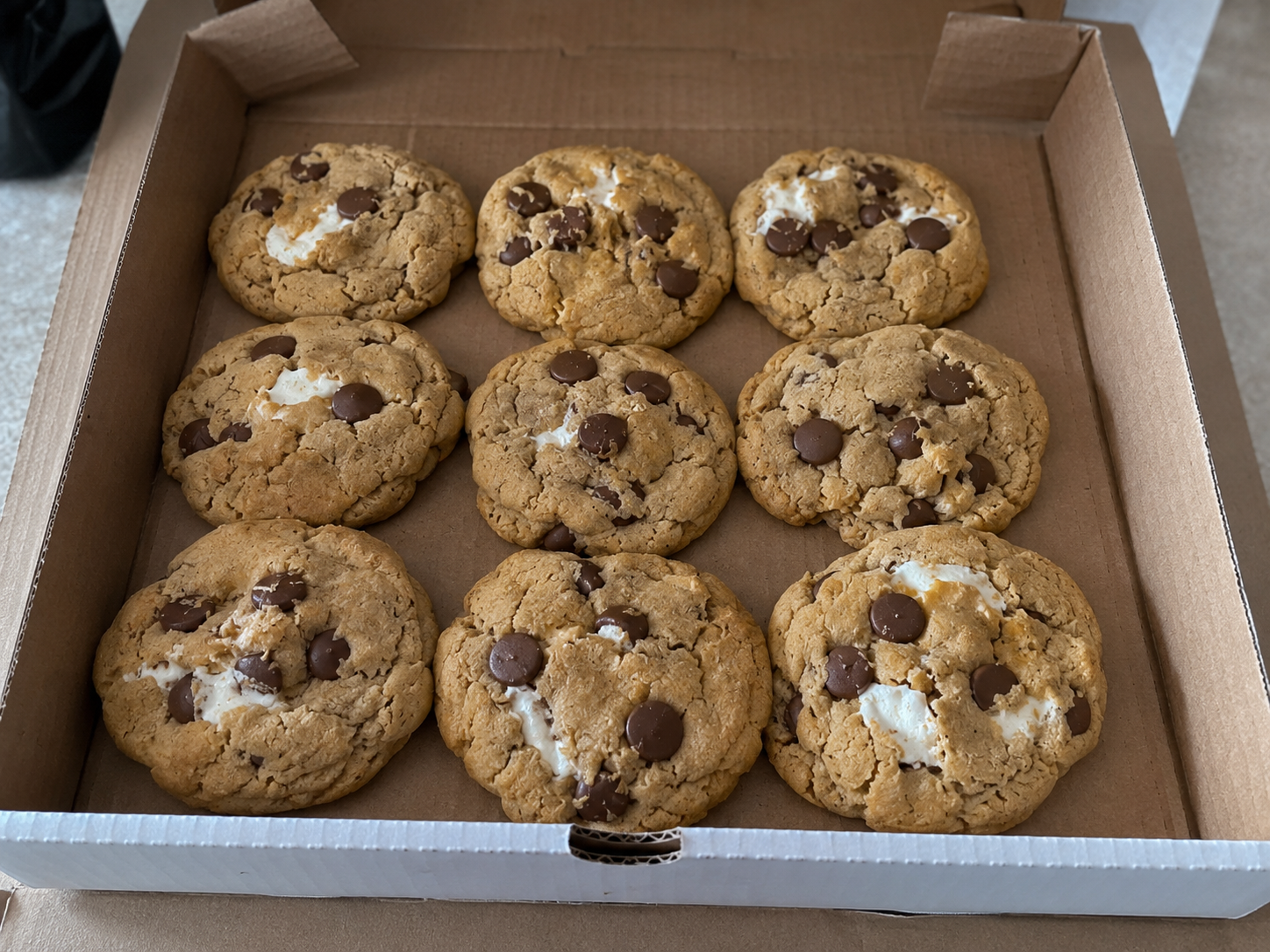 A box of ten chocolate chip cookies with chunks of white chocolate in a cardboard container.