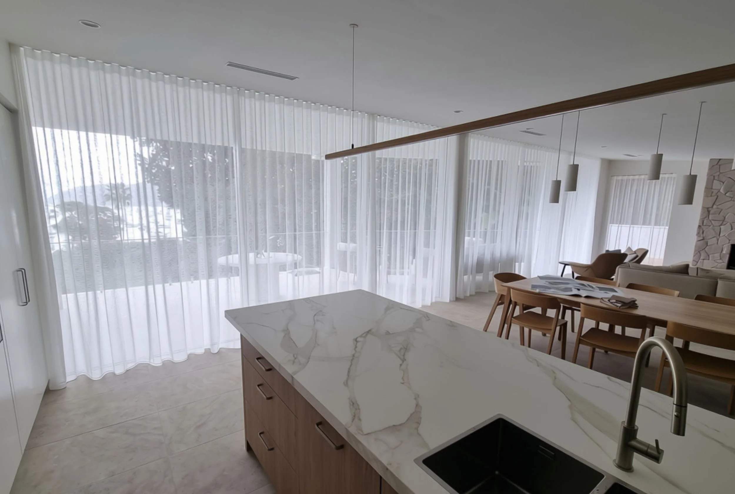 Modern open-concept kitchen with a marble countertop island, wooden dining table with chairs, large floor-to-ceiling windows with sheer curtains, and pendant lights.