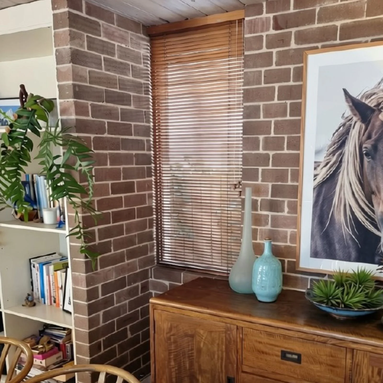 Indoor space with a brick wall, a window with blinds, a wooden table with blue vases, a framed horse portrait, a white bookshelf with books, and decorative plants.