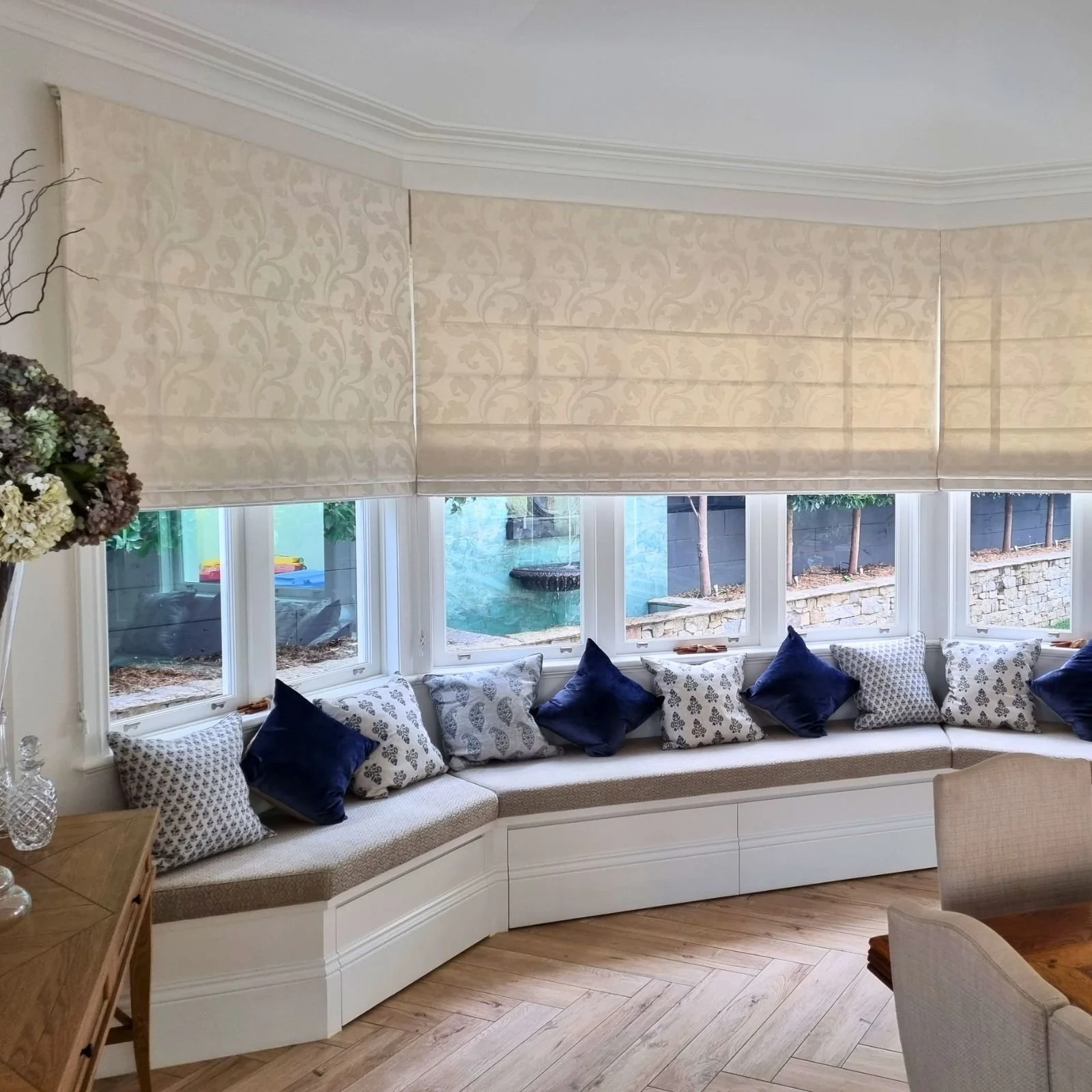 Living room with large bay window, cream-colored window seat with decorative pillows, beige roman shades, hardwood flooring, partial view of a dining area, and outdoor garden with trees and a fountain.