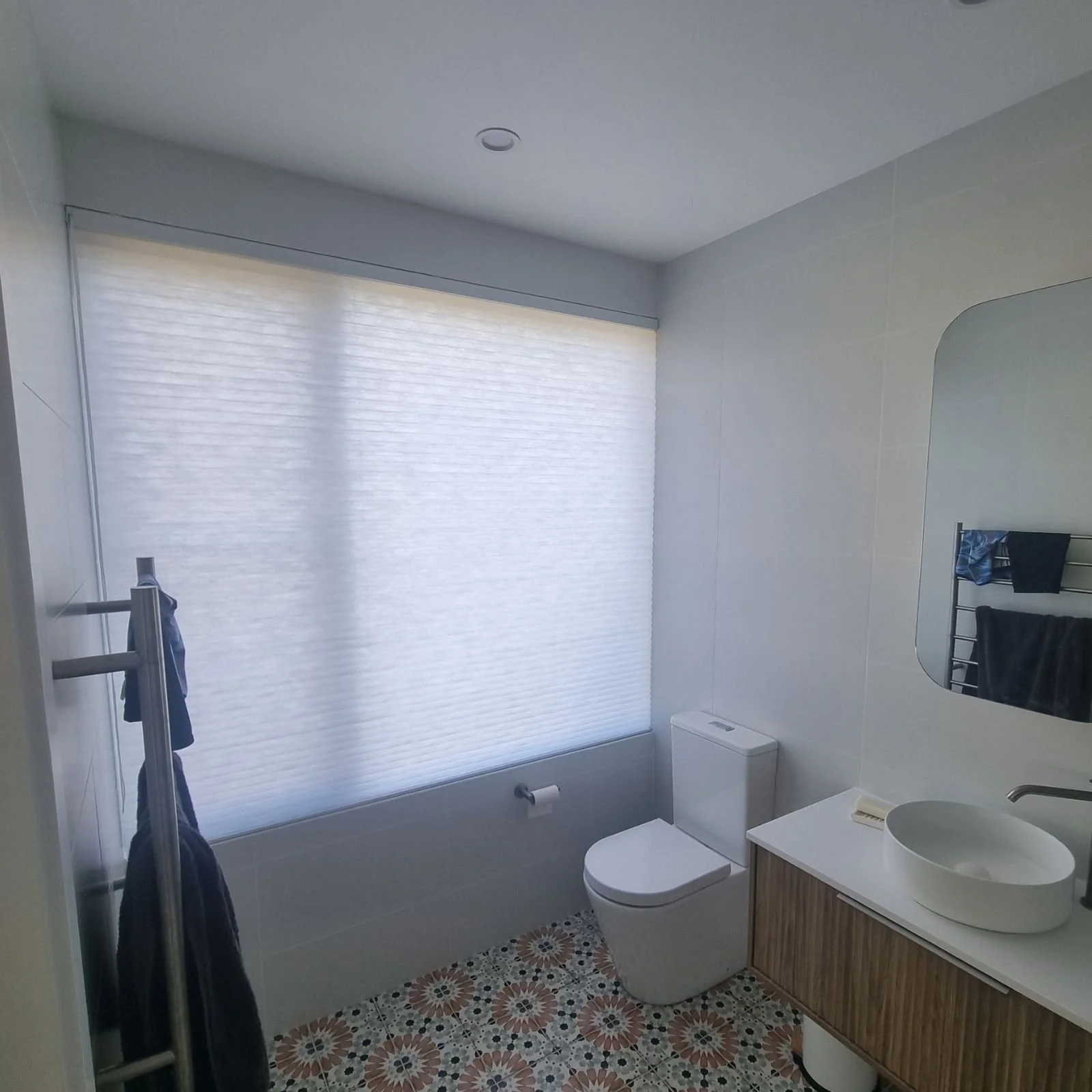 Bathroom with a window covered by a white roller blind, toilet, mirror, sink, and patterned tile floor.