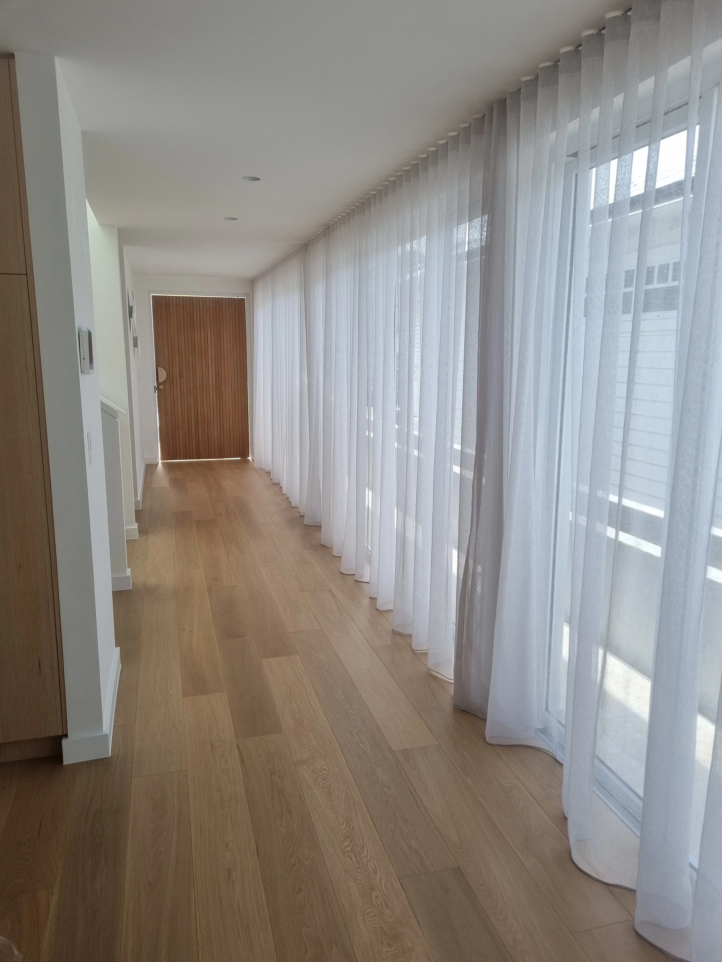 Bright hallway with wooden flooring, white walls, large window with sheer white curtains, and a wooden front door at the end.
