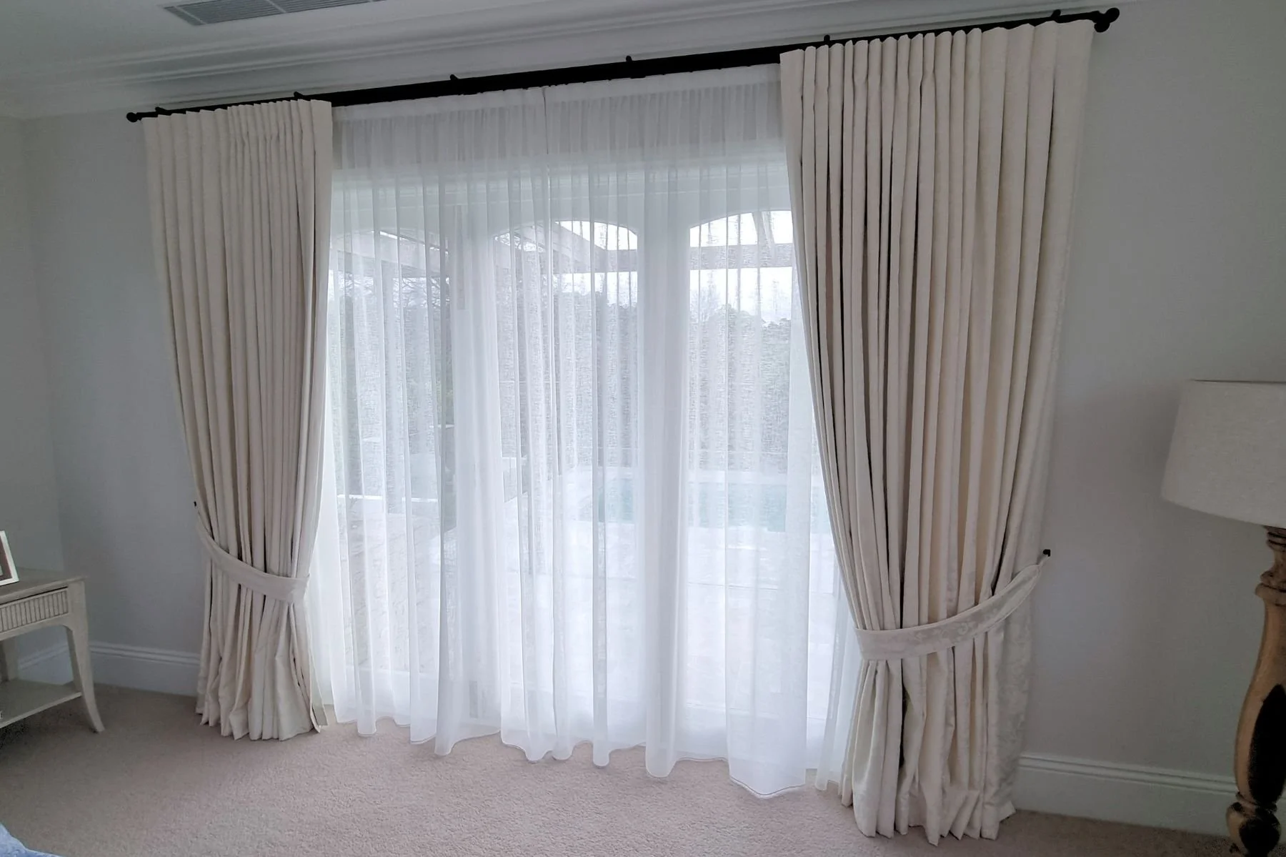 White sheer curtains with beige drapes on a large window, outside view of trees and cloudy sky.