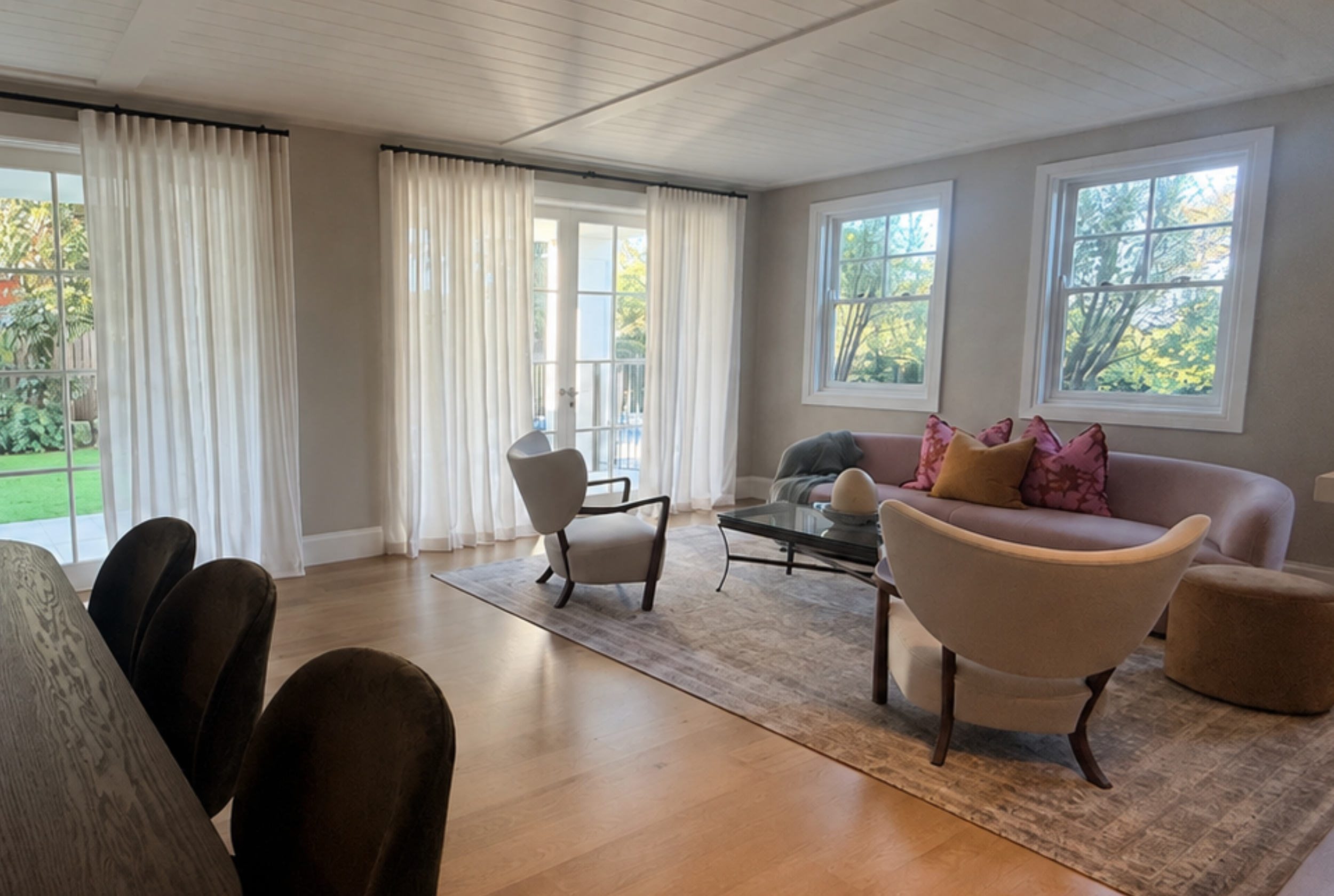 Living room with cream-colored curtains, large windows showing greenery outside, a pink sofa with pink and mustard pillows, a patterned rug, a black coffee table, and various chairs.