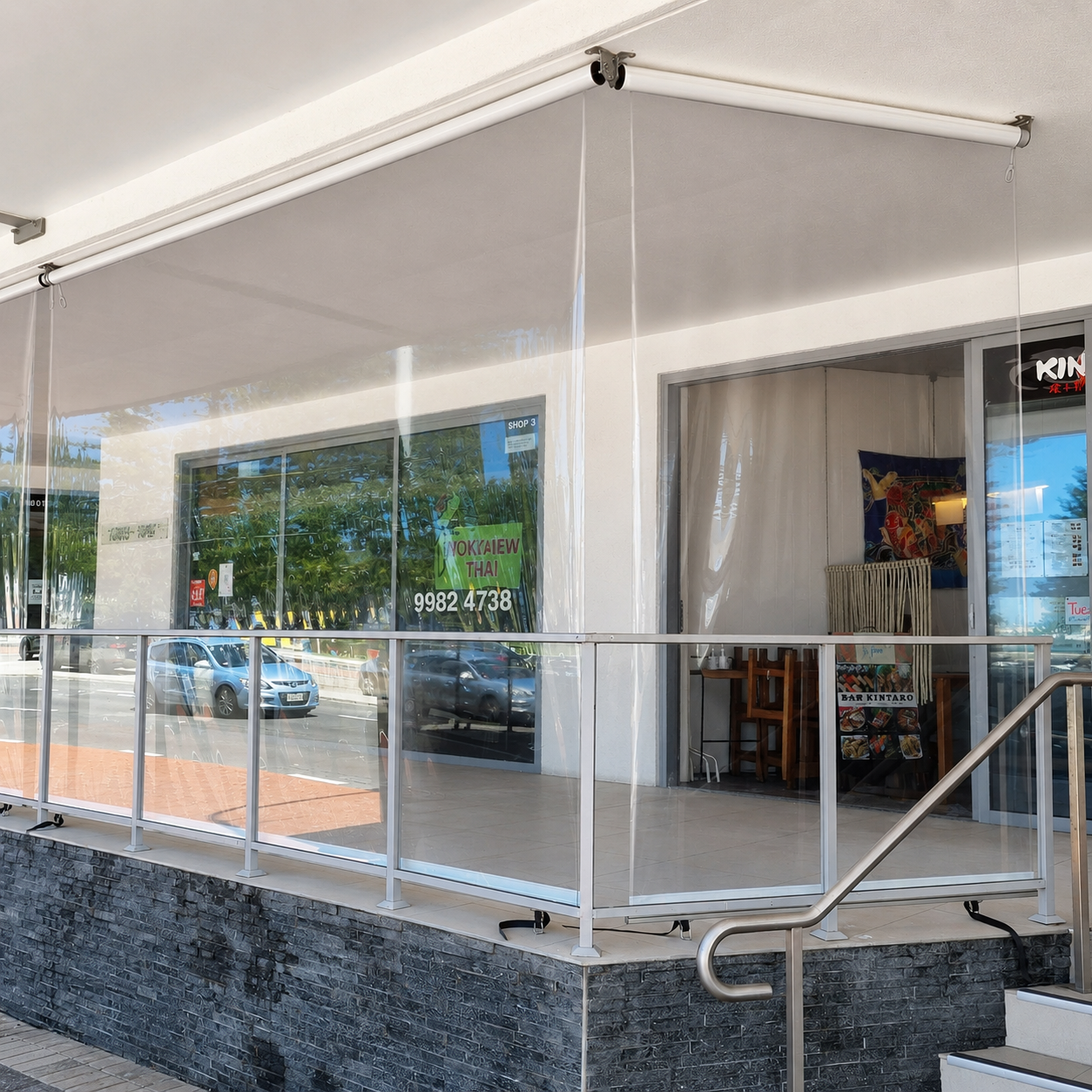 A storefront with large glass windows and a small balcony with a metal railing, displaying signs for Thai food and other businesses, with parked cars and trees visible through the windows.