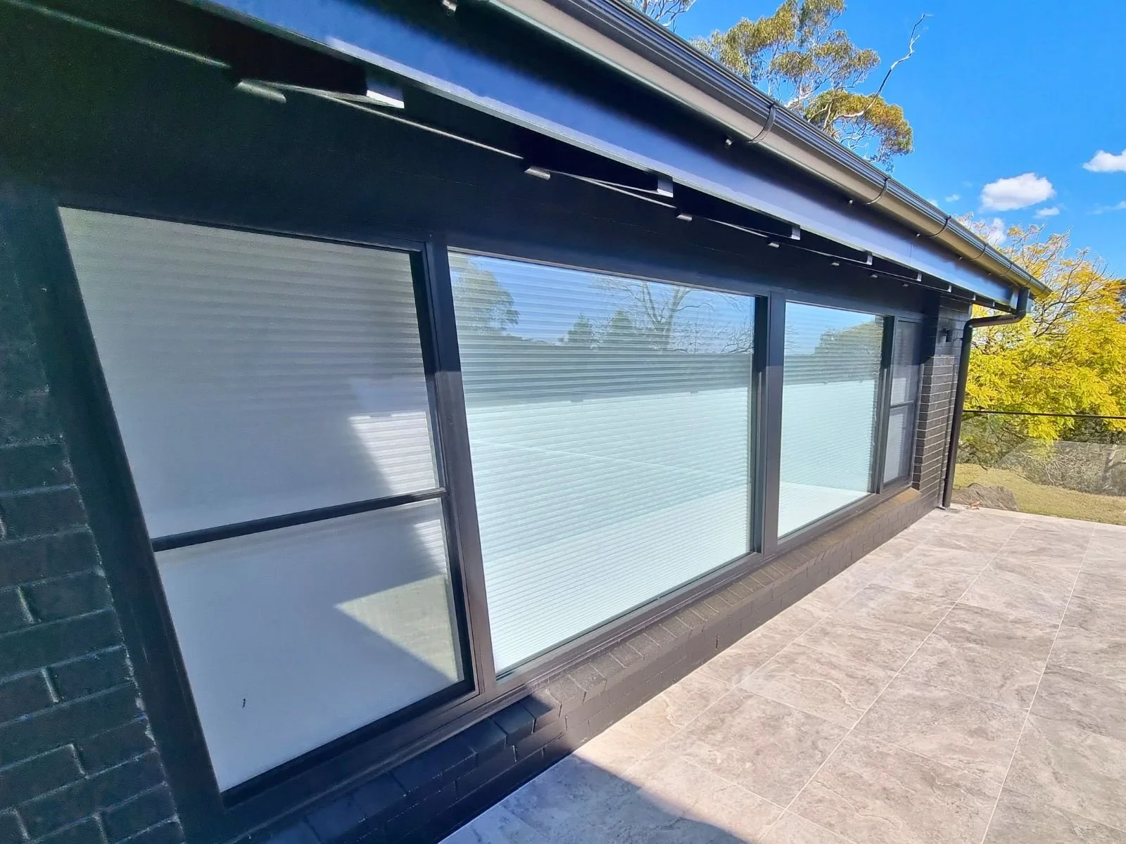View of a modern building's glass sliding doors with shading blinds, black brick exterior, and a spacious tiled balcony with a view of trees and a bright blue sky.