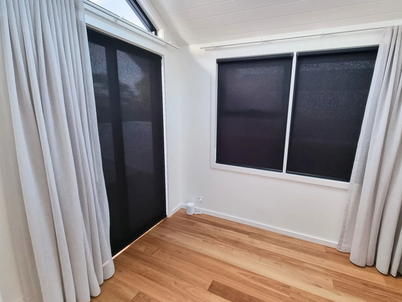 A corner of a room with light-colored curtains, black window shades, hardwood floor, and white walls. There is a white air purifier or electronic device on the floor and a white electrical outlet.