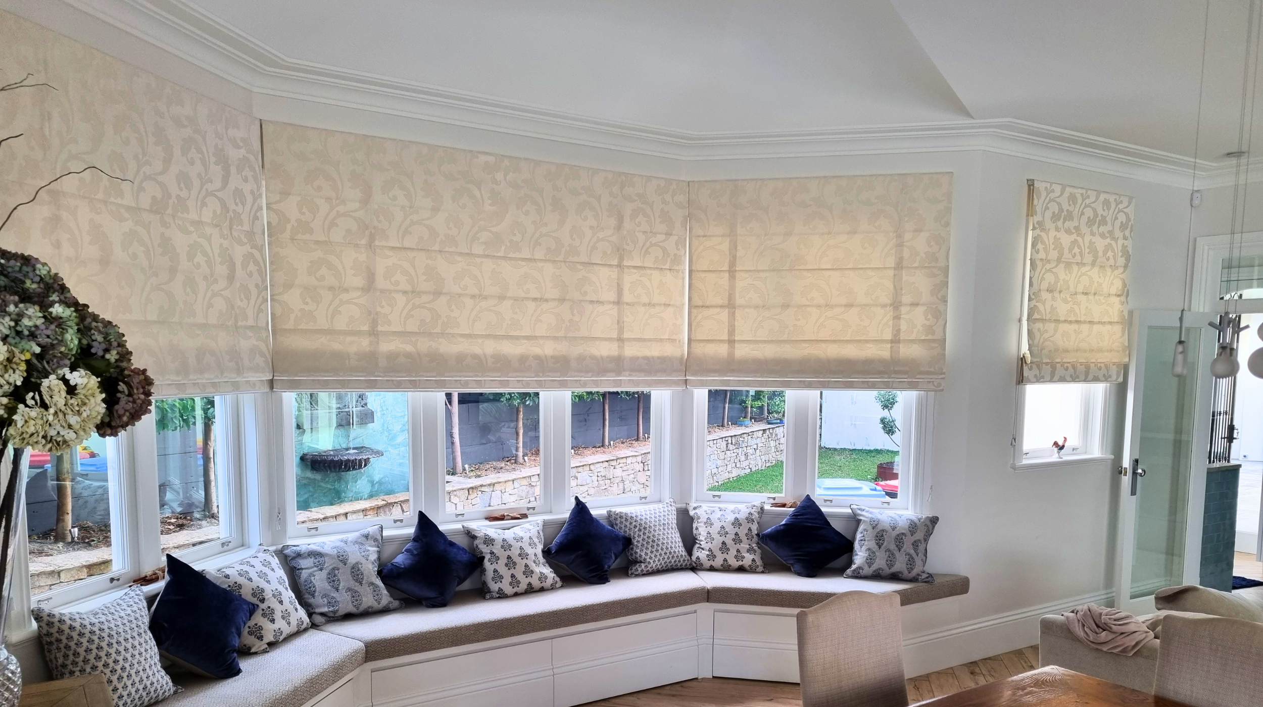 Living room with large bay windows covered by beige patterned roman shades and blue decorative pillows on a curved built-in window seat.