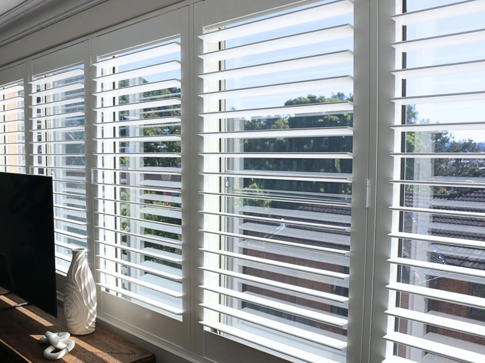 Room with large windows having white plantation shutters, outside view of trees and neighboring rooftops.