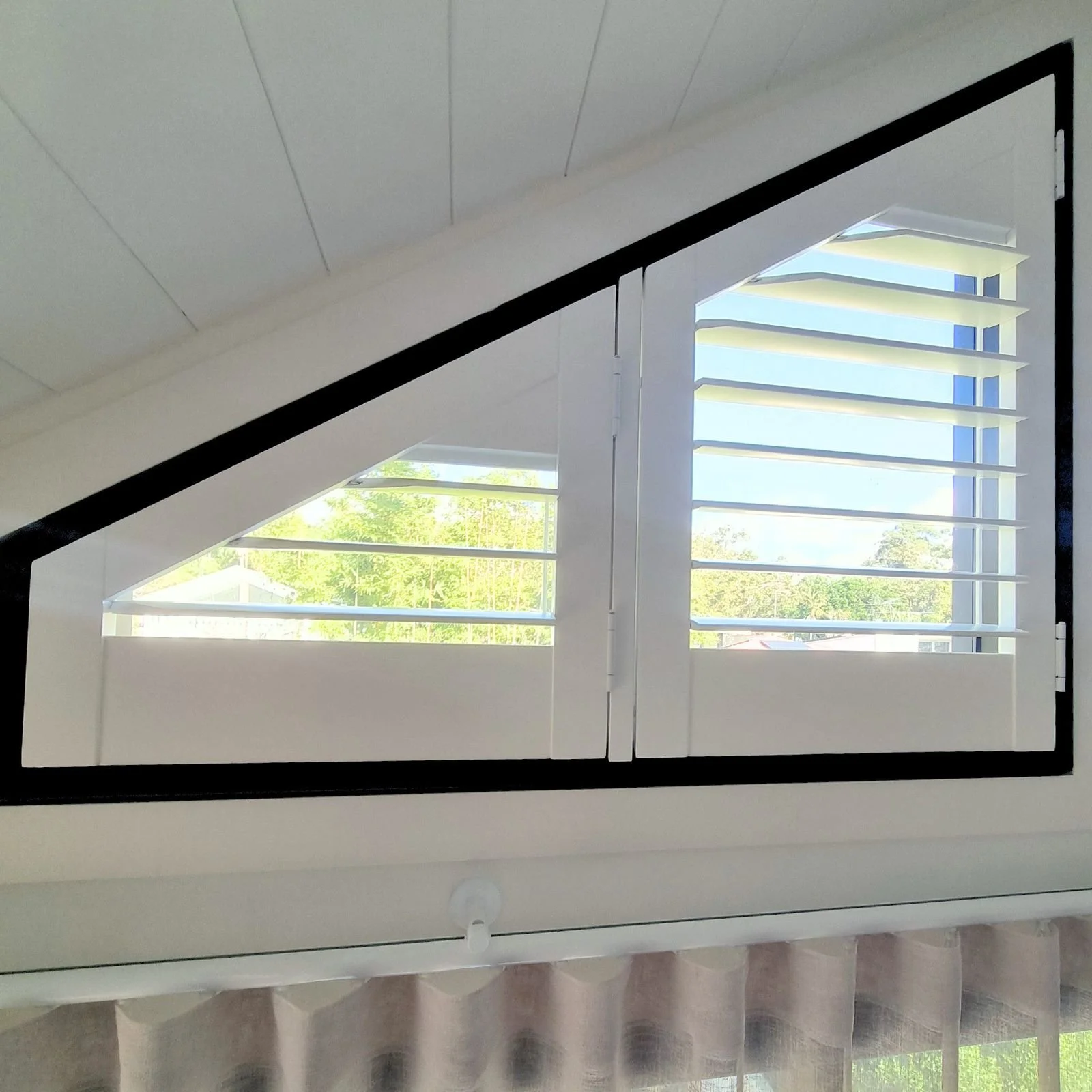 View of a room window with white blinds and a black frame, letting in sunlight and showing green trees outside.