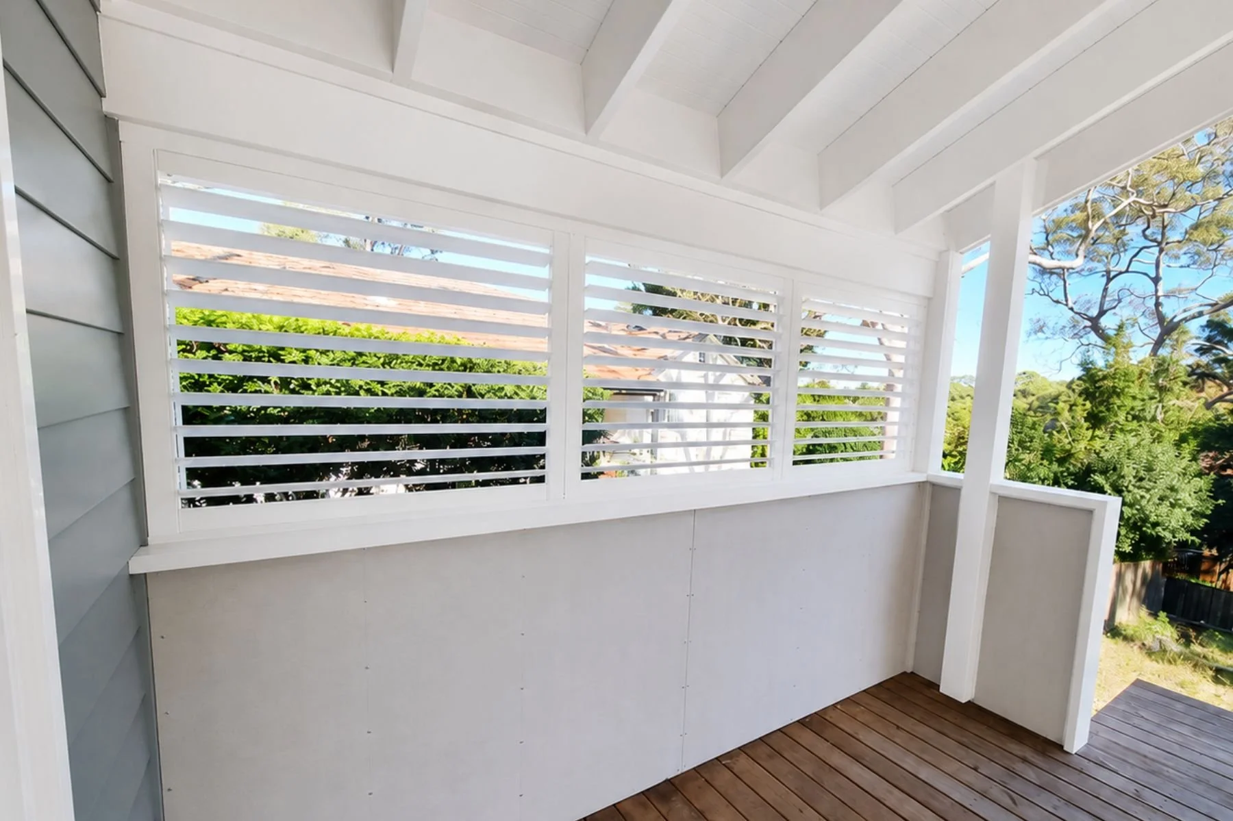 A balcony with white wooden slatted railing, gray siding wall, and wooden floor, overlooking green trees and a bright blue sky.