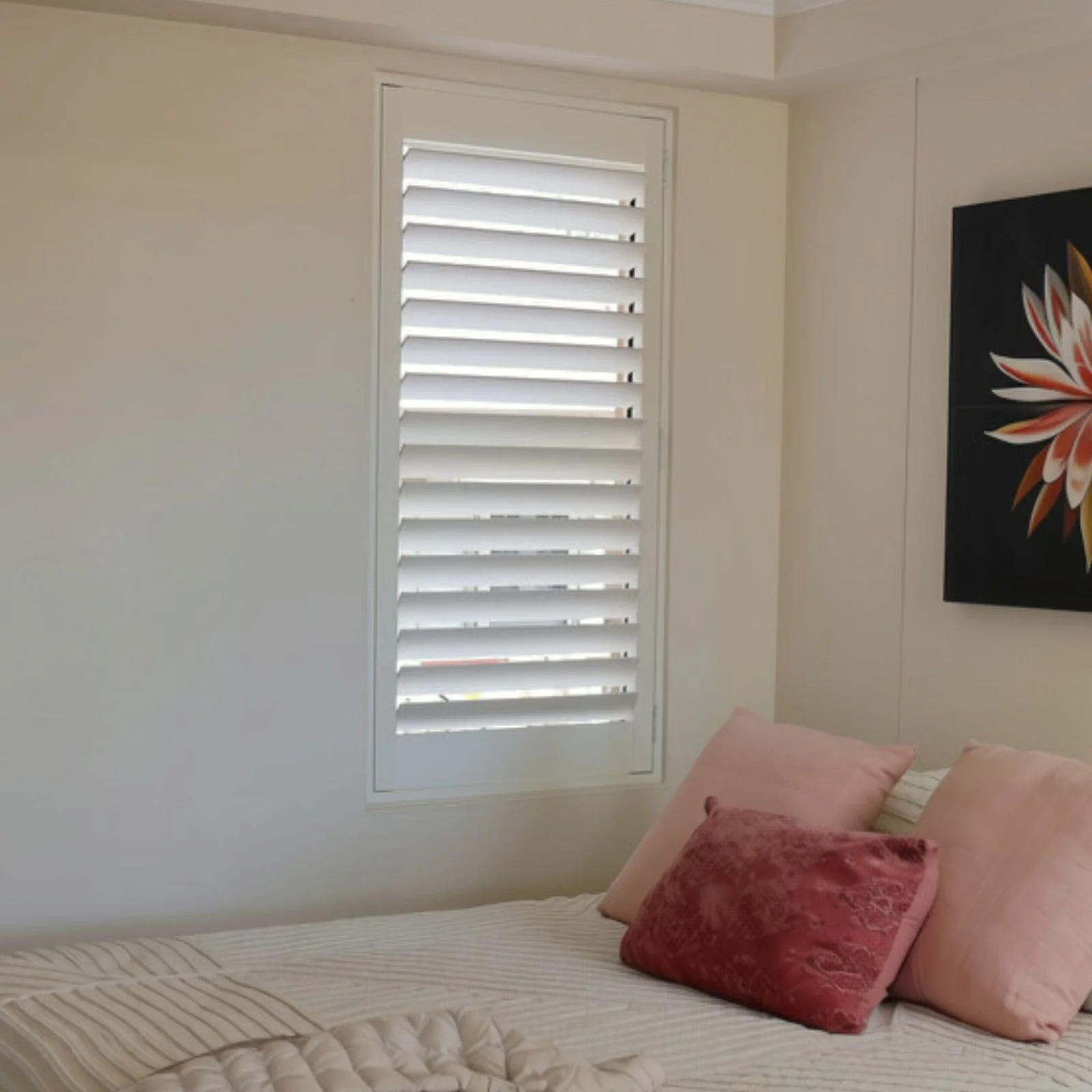 A bedroom with a window covered by white plantation shutters, a bed with a beige and white striped bedspread, and pink pillows. There is a black wall decoration with a floral design on the right side of the image.