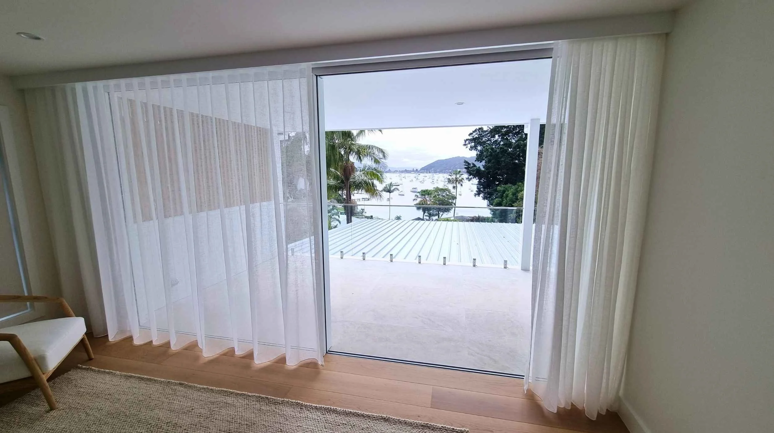 View of a balcony with glass railing overlooking a marina with boats, palm trees, and distant mountains through sliding glass doors decorated with sheer white curtains inside a modern apartment.