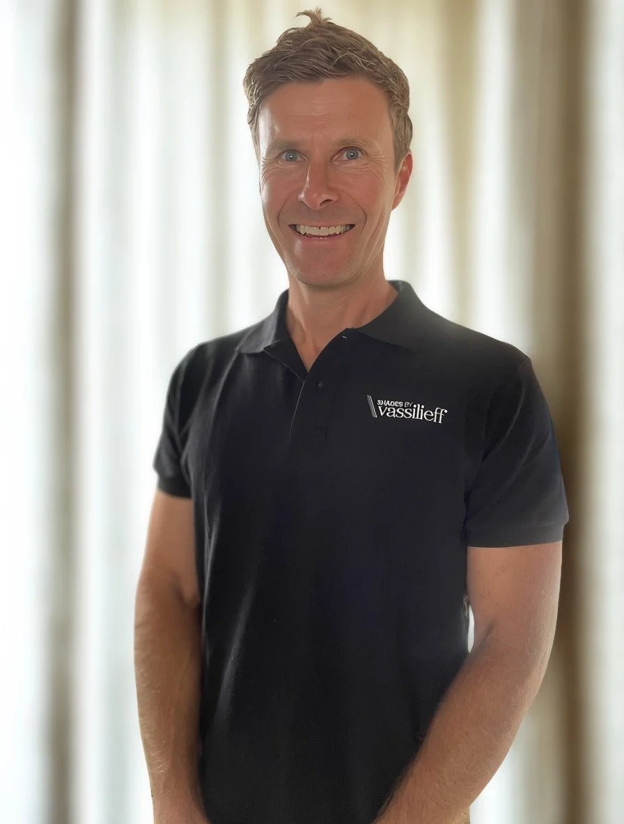 Darren Vassilieff wearing a black polo shirt with the logo and text 'SHADES BY Vassilieff' on it, standing in front of curtains.