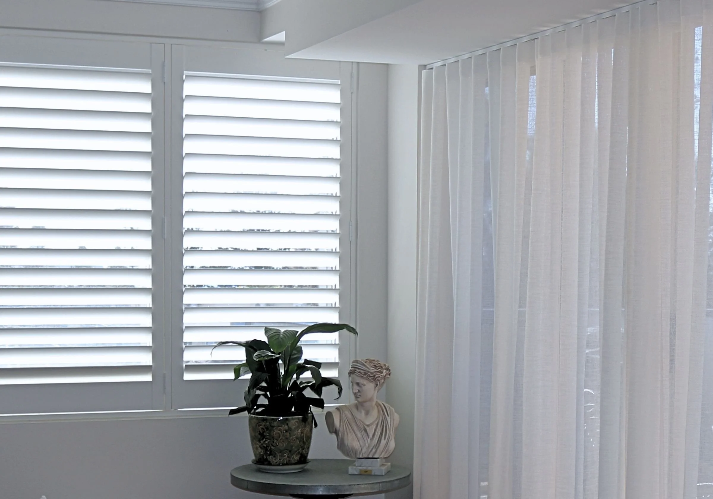 A white window with plantation shutters on the left and sheer curtains on the right. In front, a table holds a potted plant, a bust sculpture, and a small decorative object.