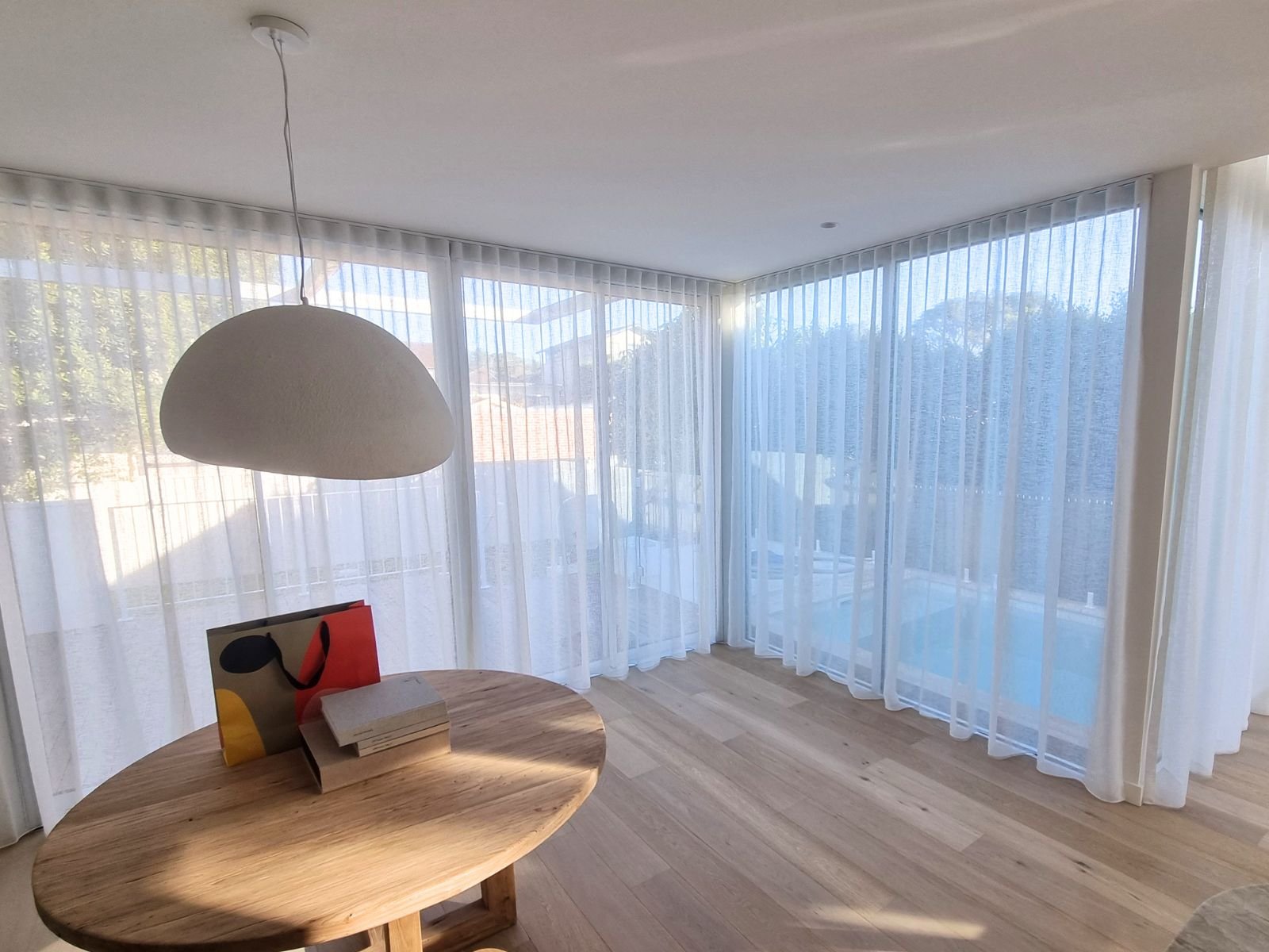 Interior of a room with large windows covered with sheer white curtains, a round wooden table with books and a colorful bag on it, and a large hanging white lamp.