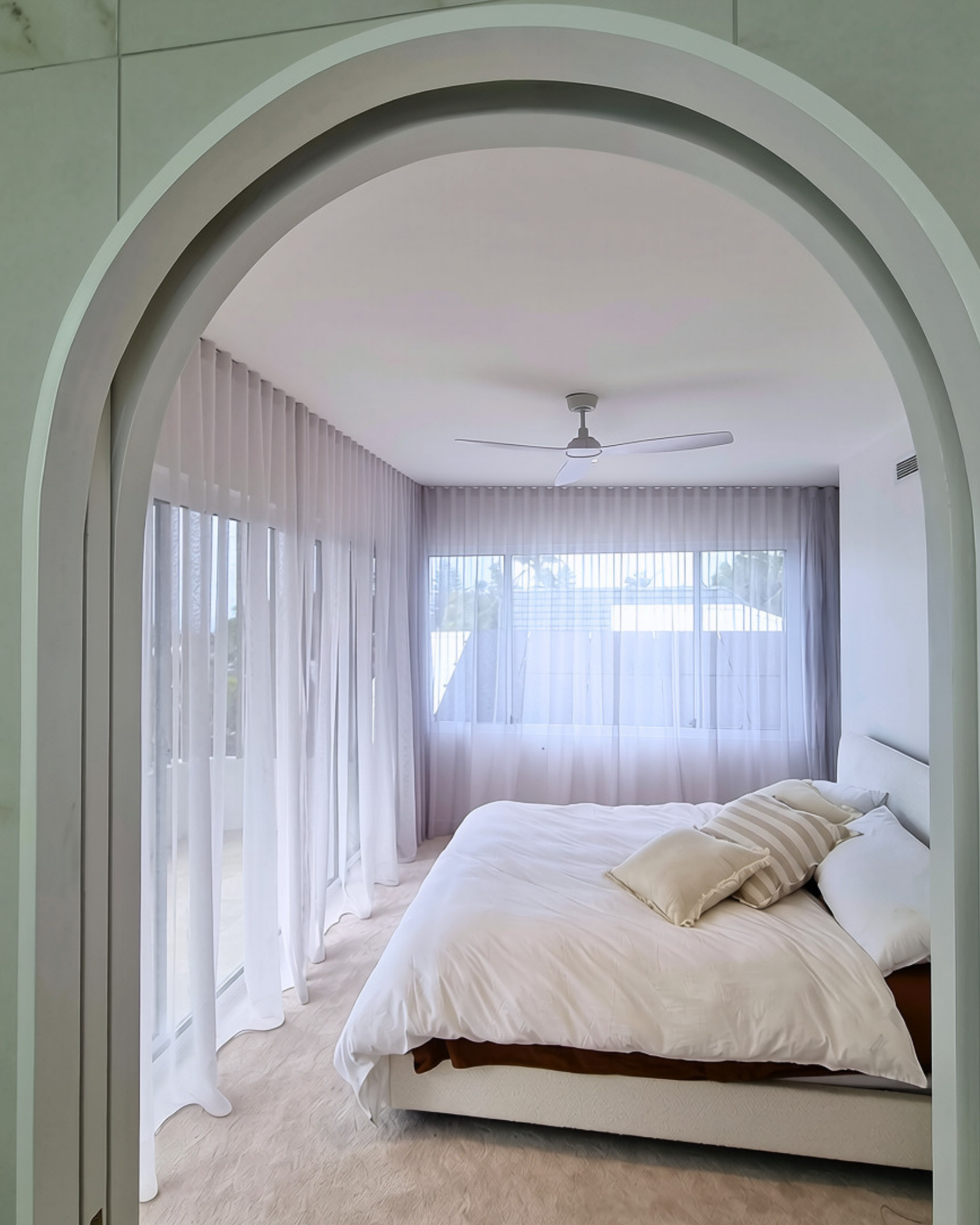 A bedroom with a large bed with white sheets, a striped pillow, and a beige pillow, in front of a window with sheer white curtains. The room has a ceiling fan and a view of outdoors through the windows.