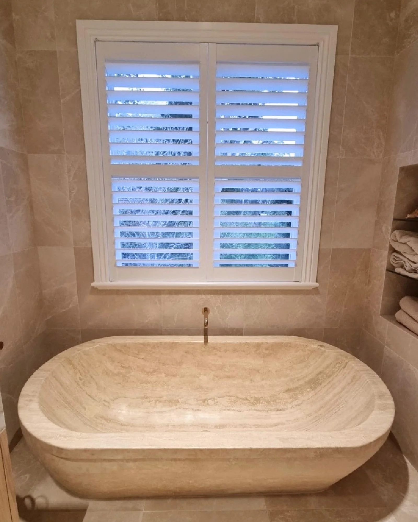 A bathroom with a built-in oval stone bathtub, surrounded by beige tiled walls, and a large window with white shutters above the tub.