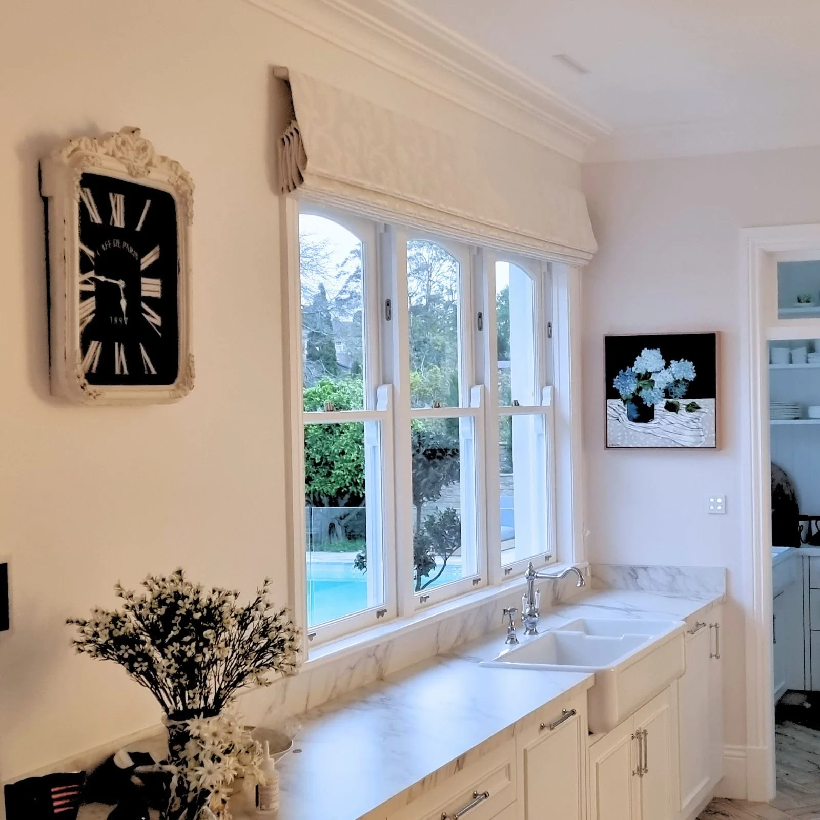 A bright kitchen with large windows and white cabinetry. A black and white floral painting hangs on the wall, and a bouquet of white flowers is on the marble countertop near a white farmhouse sink with a chrome faucet. A vintage-style wall clock is on the left wall.